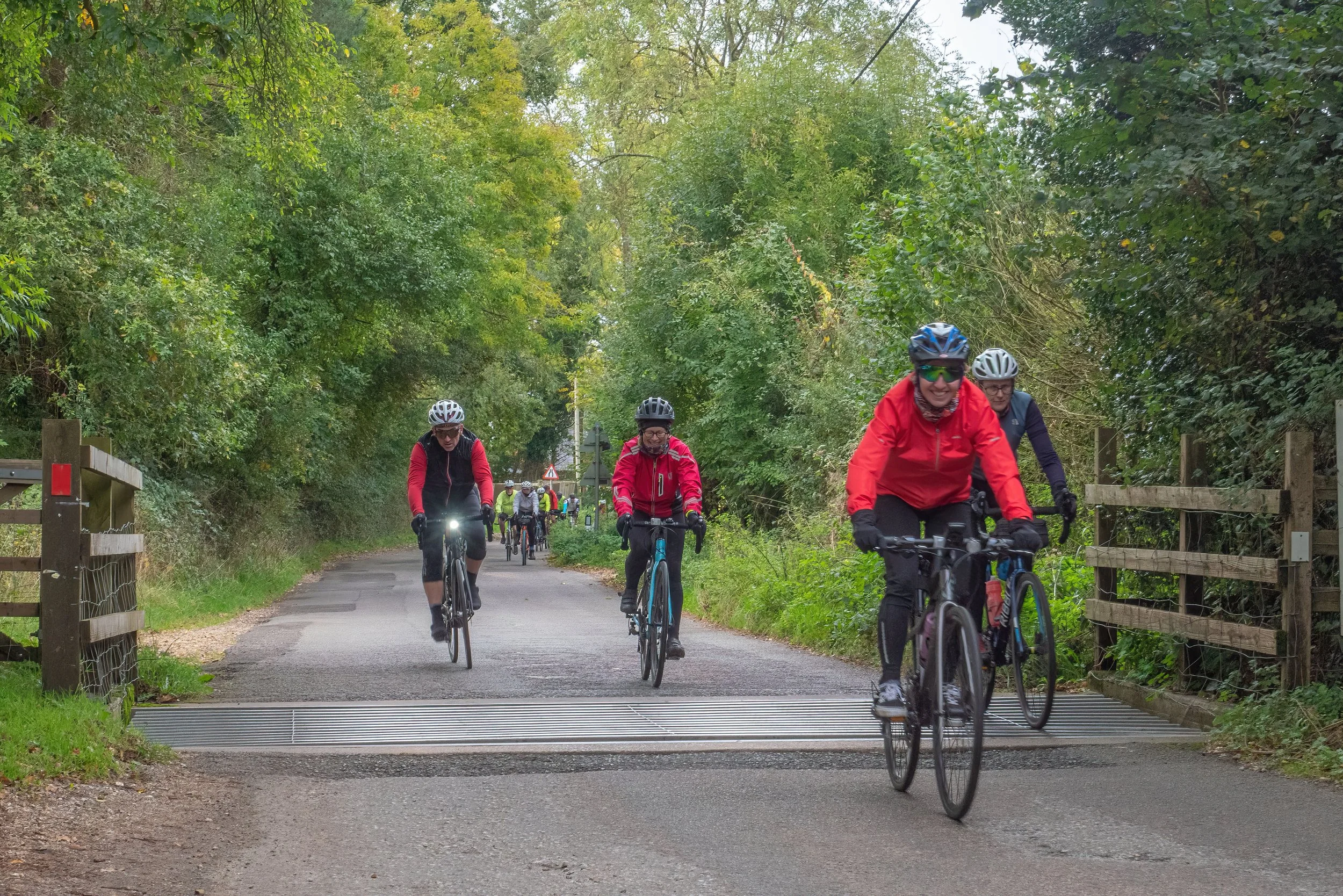 Group of people riding bicycles on a shaded path through greenery, wearing helmets and outdoor clothing.