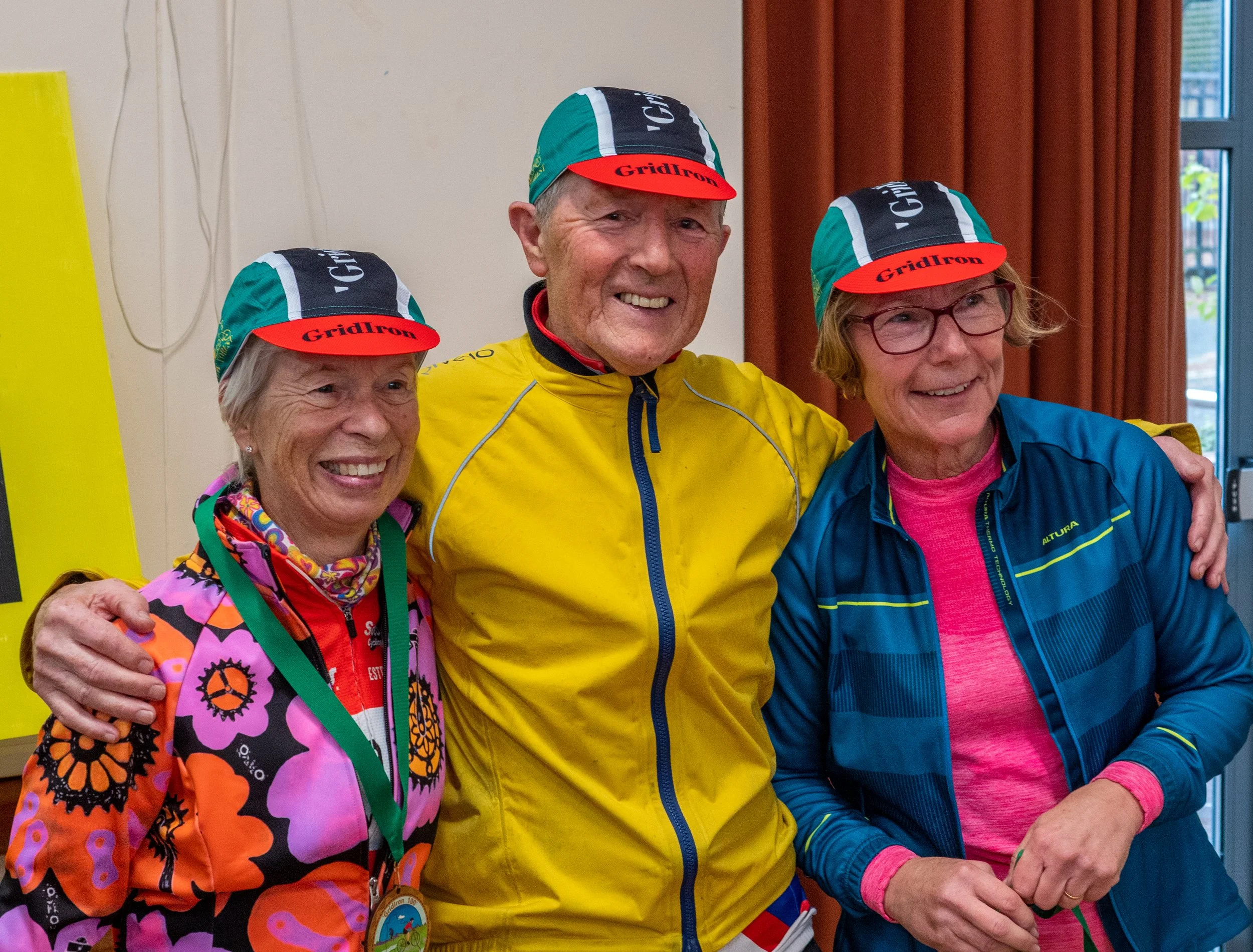 Three cheerful elderly individuals, two women and one man, wearing colorful cycling jerseys and hats with a logo "GridIron." They are standing close together, smiling, and appear to be at an indoor event, possibly celebrating or participating in a cy