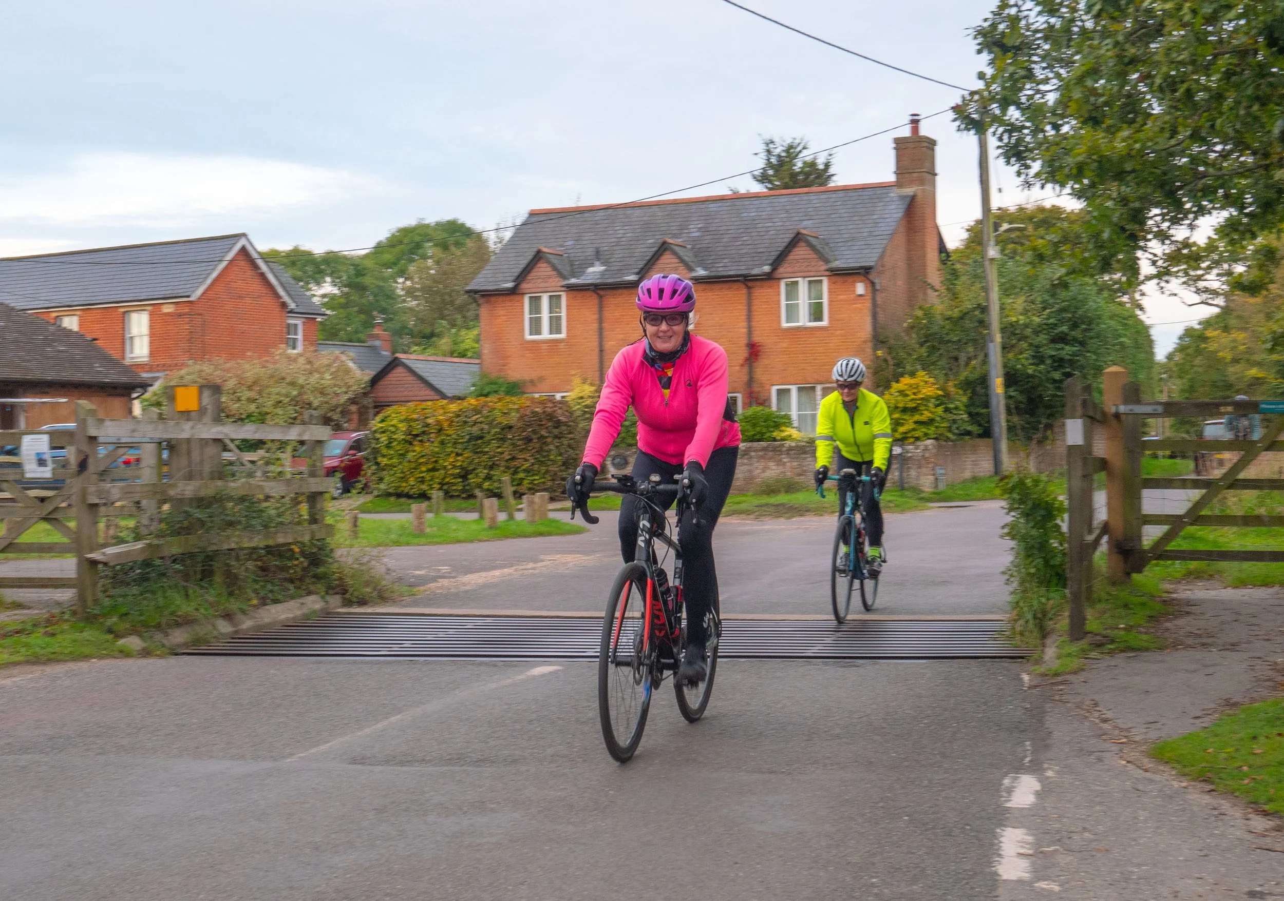 Two women riding bicycles on a suburban street, wearing helmets with one in a pink jacket and the other in a yellow jacket, with brick houses and trees in the background.