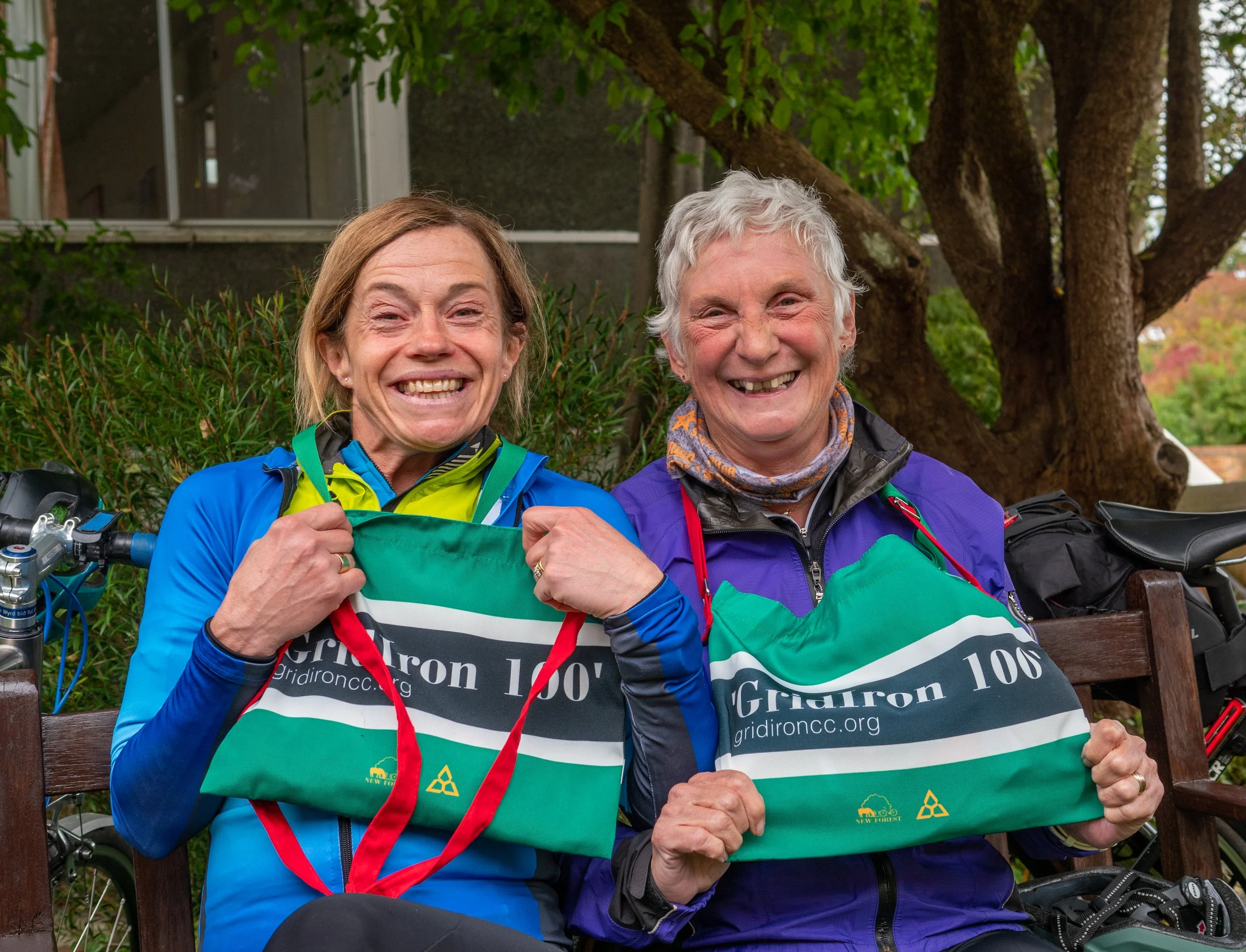 Two smiling women sitting on a park bench, wearing colorful outdoor gear and holding bags with the logo 'Gridiron 100' and a website 'gridironcc.org,' with green trees and a building in the background.