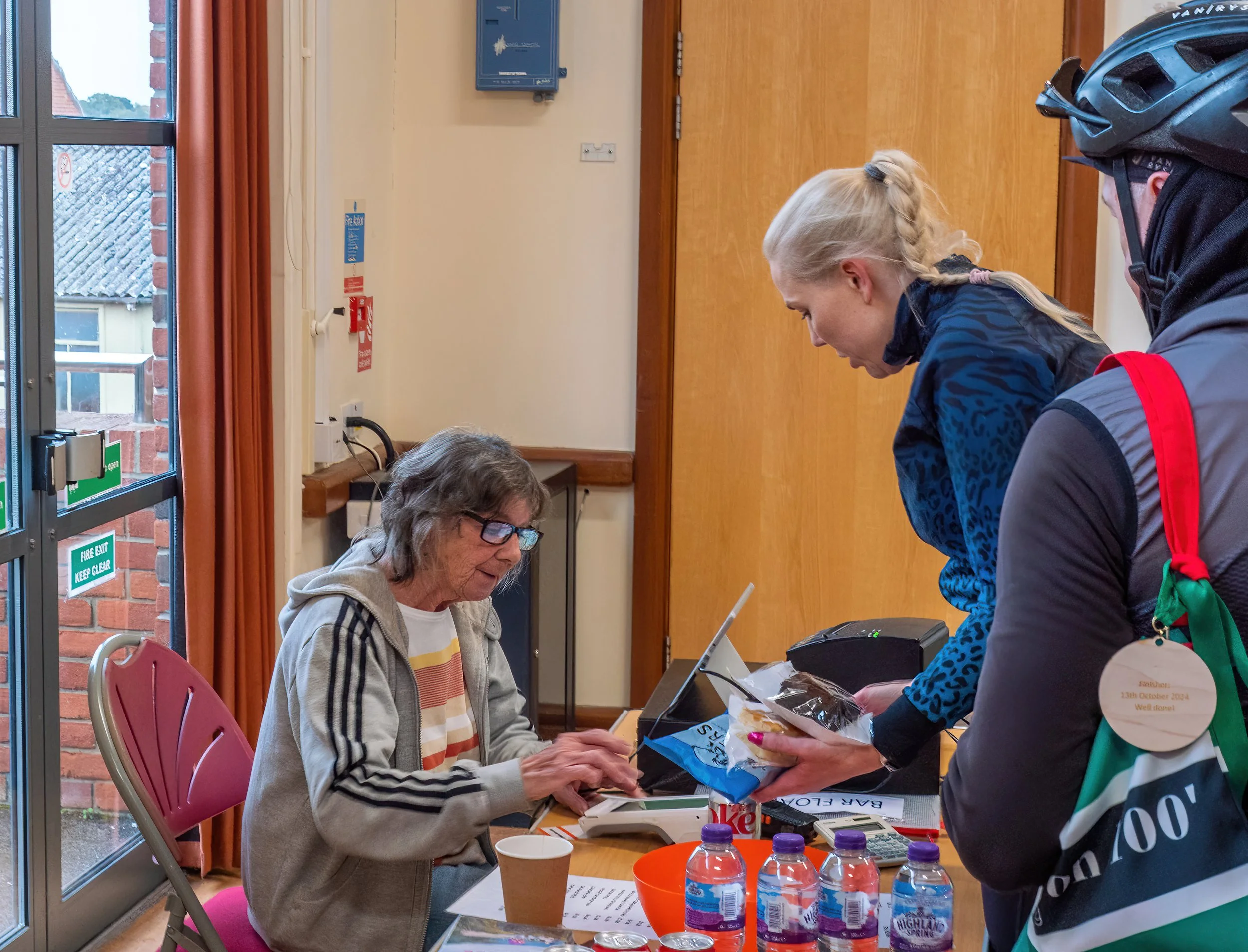 An elderly woman sitting at a table signing a paper as she receives a gift bag from a woman standing beside her. Several water bottles and a cup are on the table. The setting appears to be indoors near a door with glass panels.
