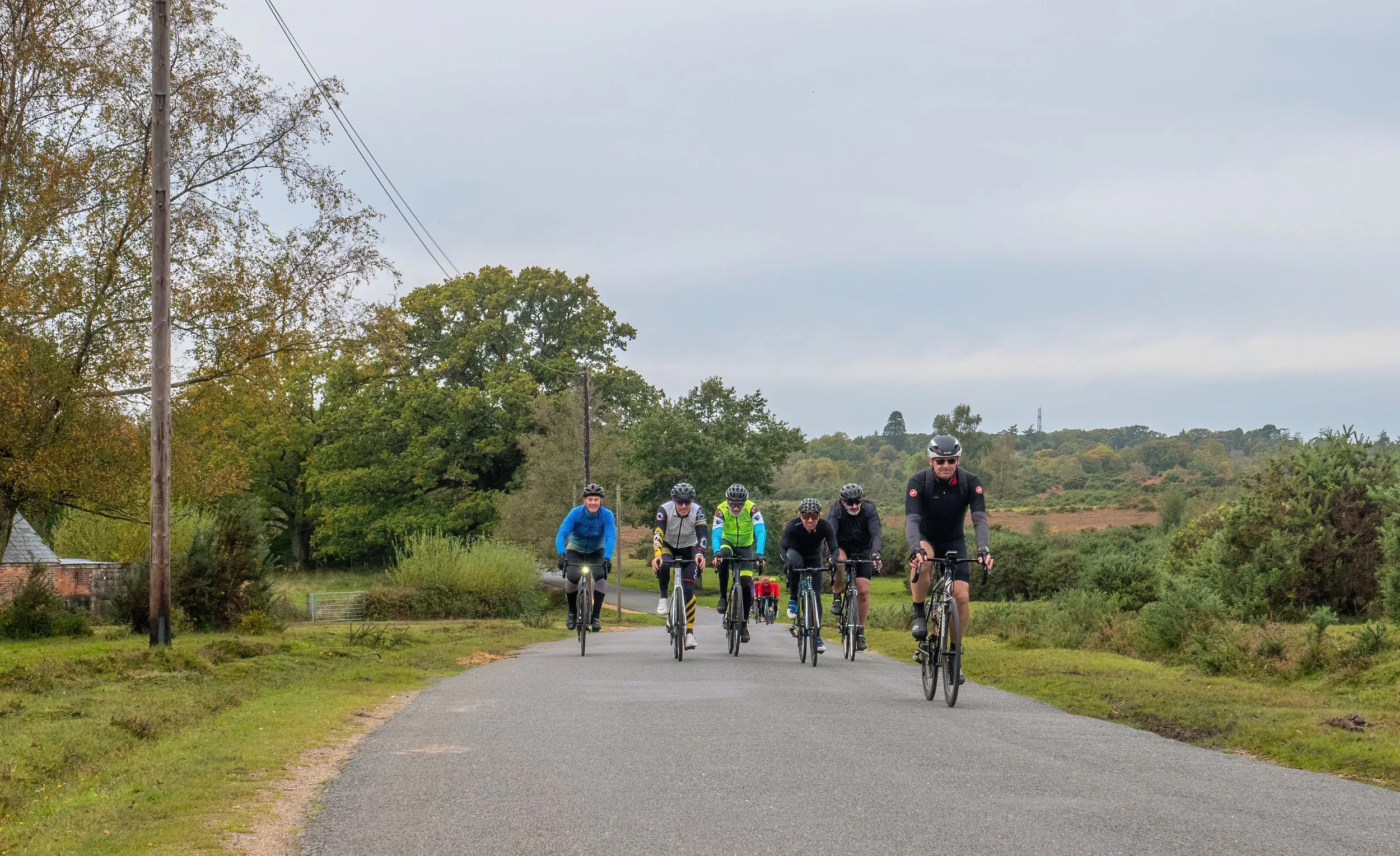 Group of cyclists riding on a rural paved road surrounded by trees and greenery, with a cloudy sky overhead.
