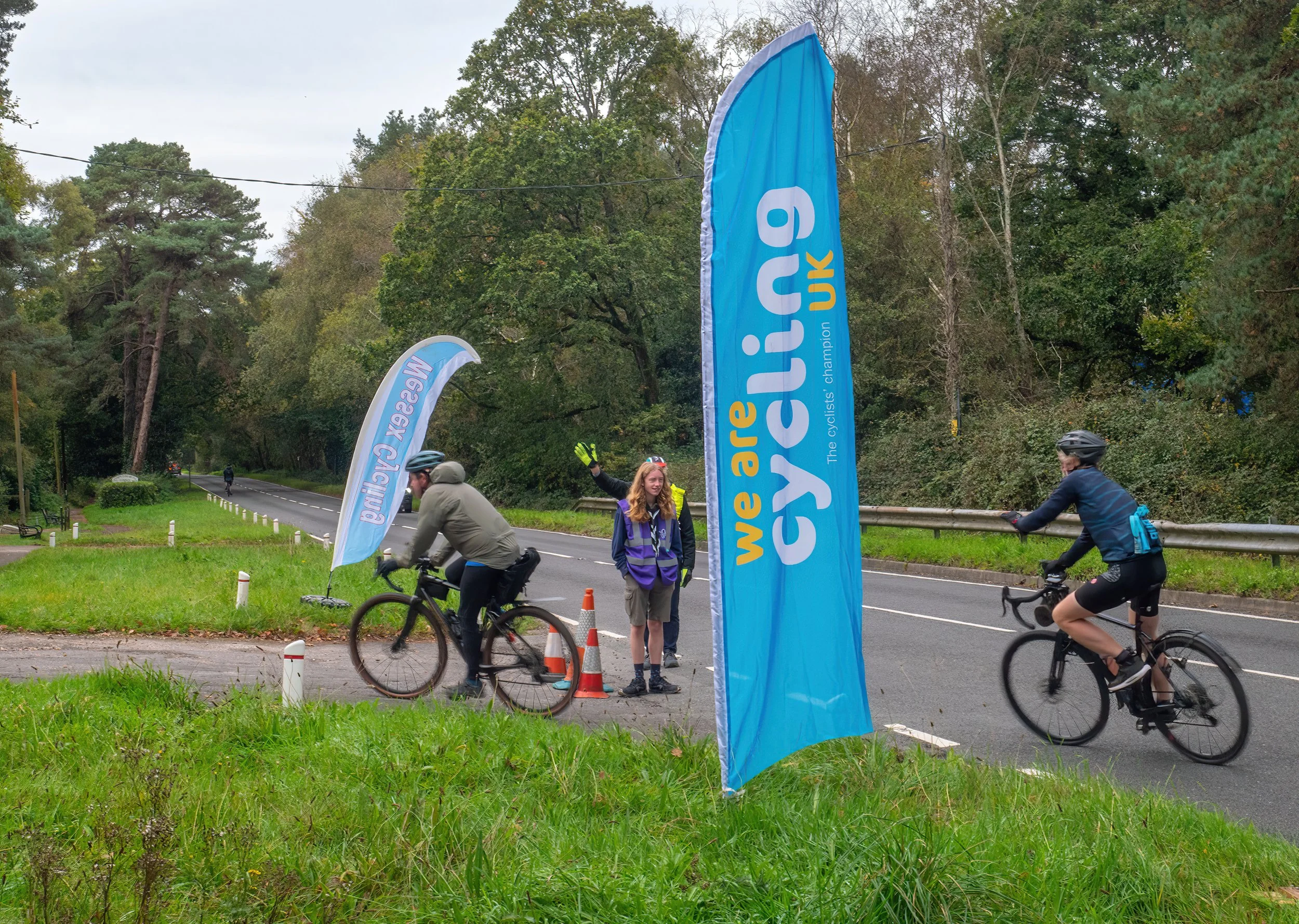 Cyclists biking past a large blue flag with yellow and white text that reads 'We are all cyclist's champion.' Two people are standing on the sidewalk, one of whom is smiling and waving at the camera, while another person is riding a bike along the ro