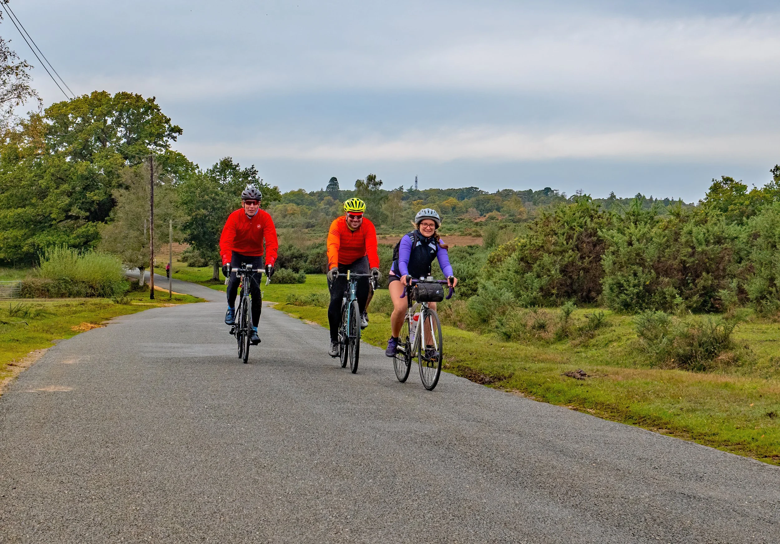 Three people riding bicycles on a paved trail in a scenic outdoor area with trees and bushes, wearing helmets and colorful outdoor clothing on a cloudy day.