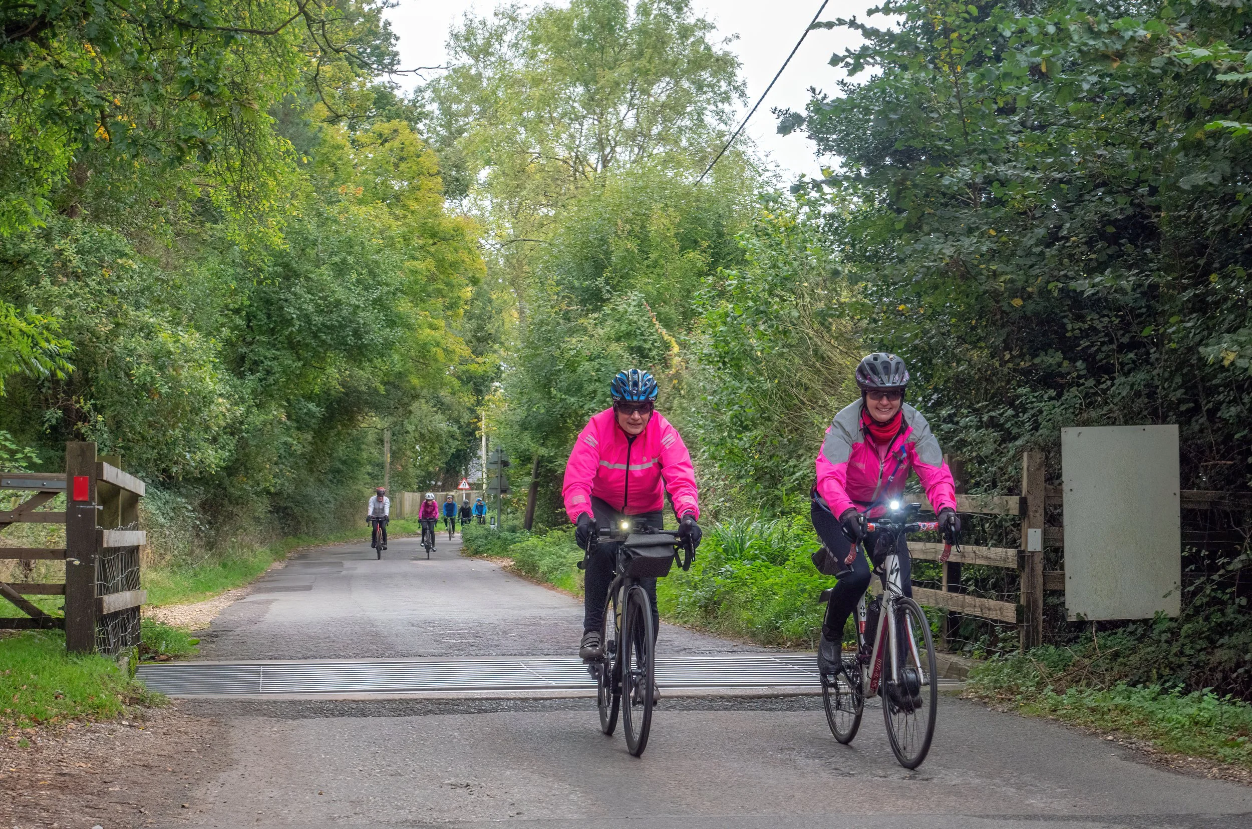 Three women riding bicycles along a tree-lined path on a cloudy day, with more cyclists following behind.