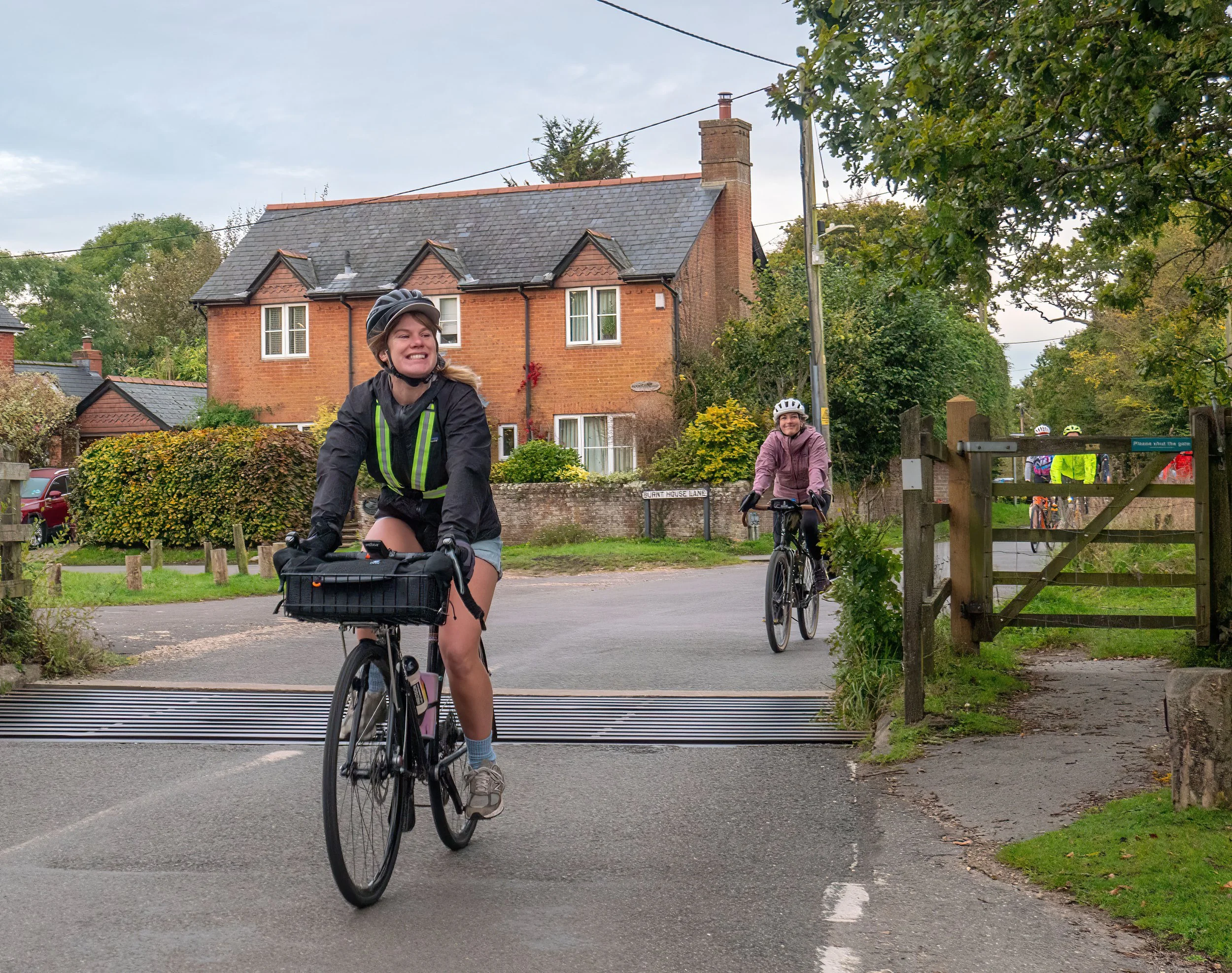 Two women riding bicycles along a suburban street with houses, trees, and a wooden gate in the background. One woman in the foreground is wearing a helmet and smiling.