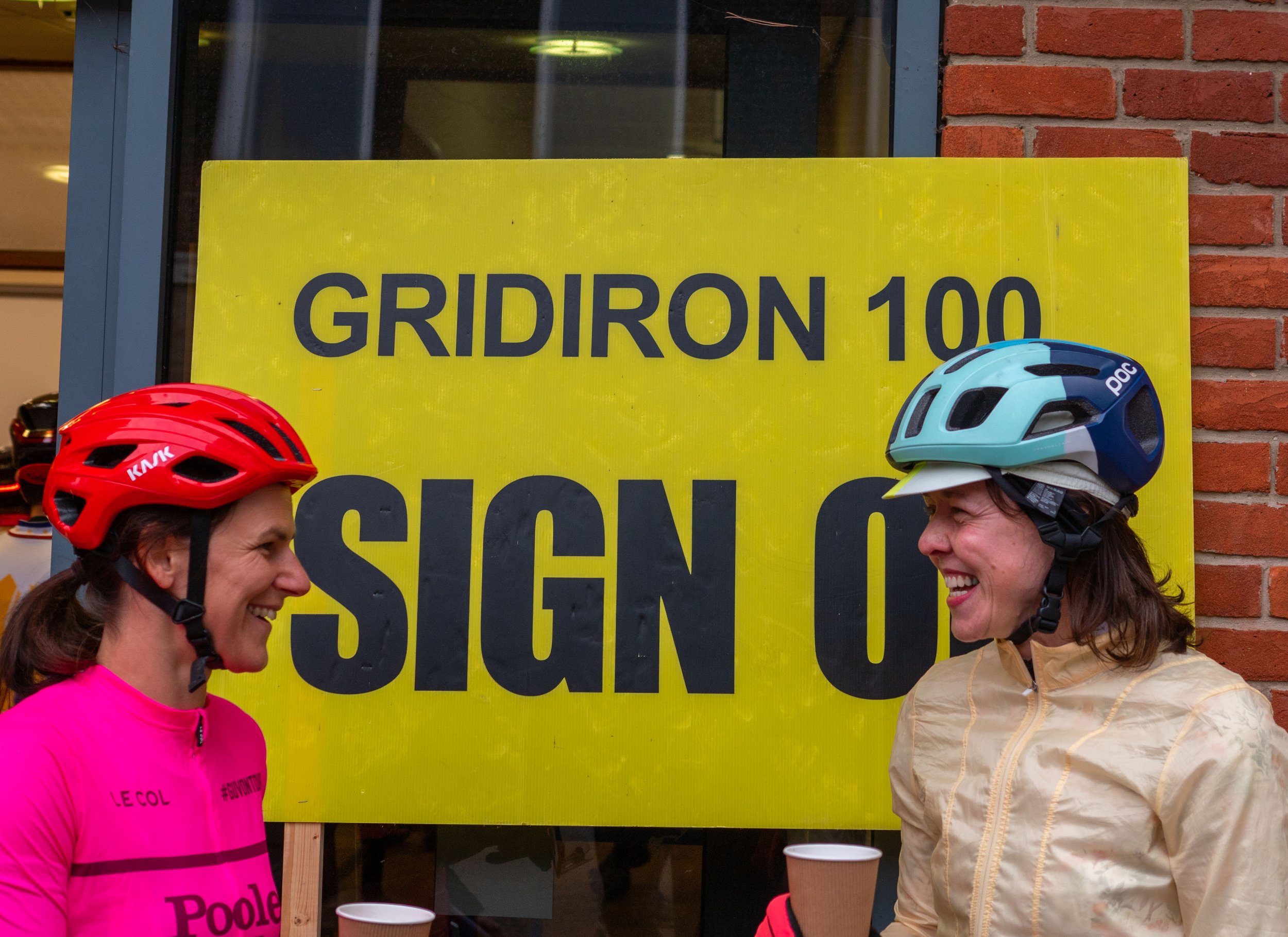 Two women wearing bicycle helmets smiling and talking outside near a building with a yellow sign that reads 'GRIDIRON 100' in the background.