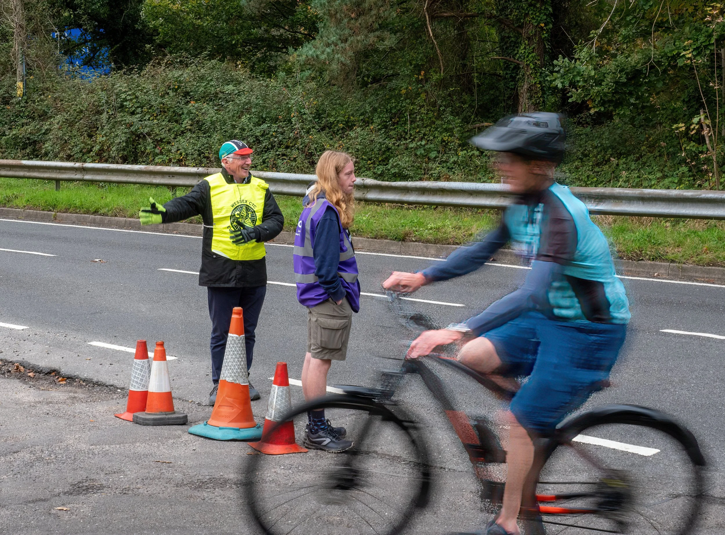 A man wearing a yellow vest and glasses standing on a street next to a row of orange traffic cones, talking with a young woman with long hair in a purple jacket and backpack. A cyclist in motion, blurred, wearing a helmet and blue jacket, rides past 