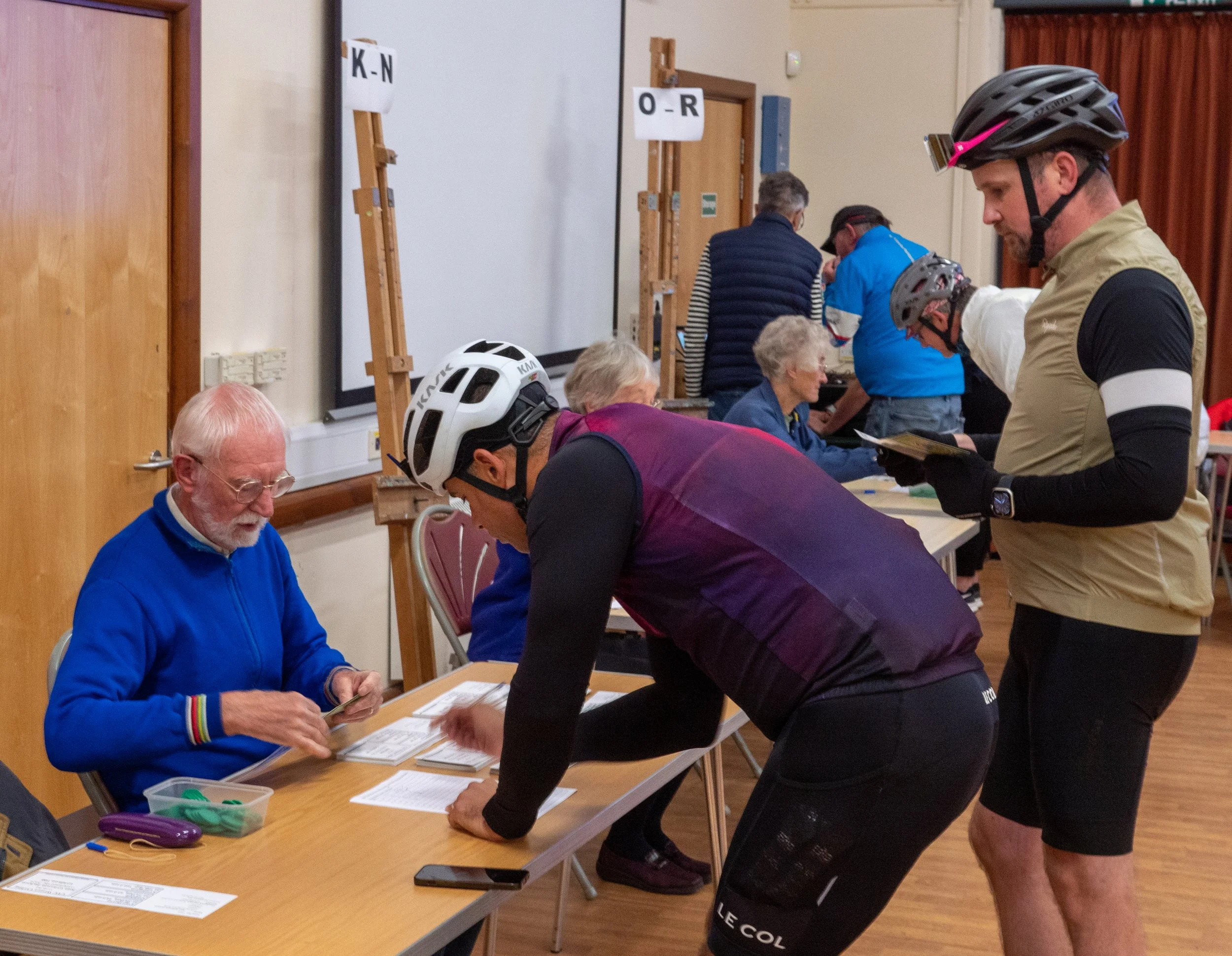 People at a registration table with signs indicating alphabetical groups, some wearing bike helmets, in an indoor setting with wooden accents and curtains.