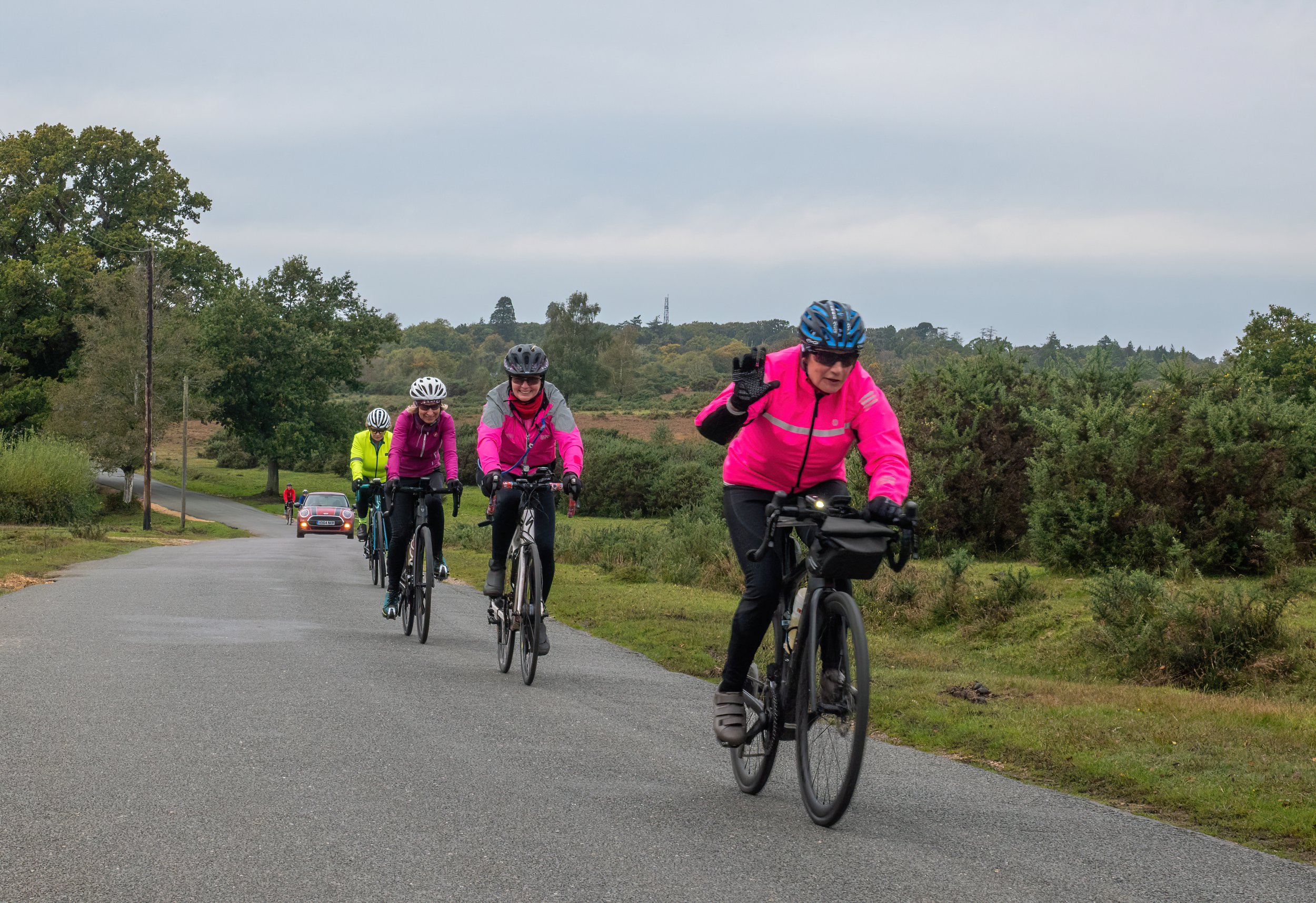 A group of people riding bicycles on a rural road, wearing helmets and colorful outdoor clothing, with trees and hills in the background under an overcast sky.