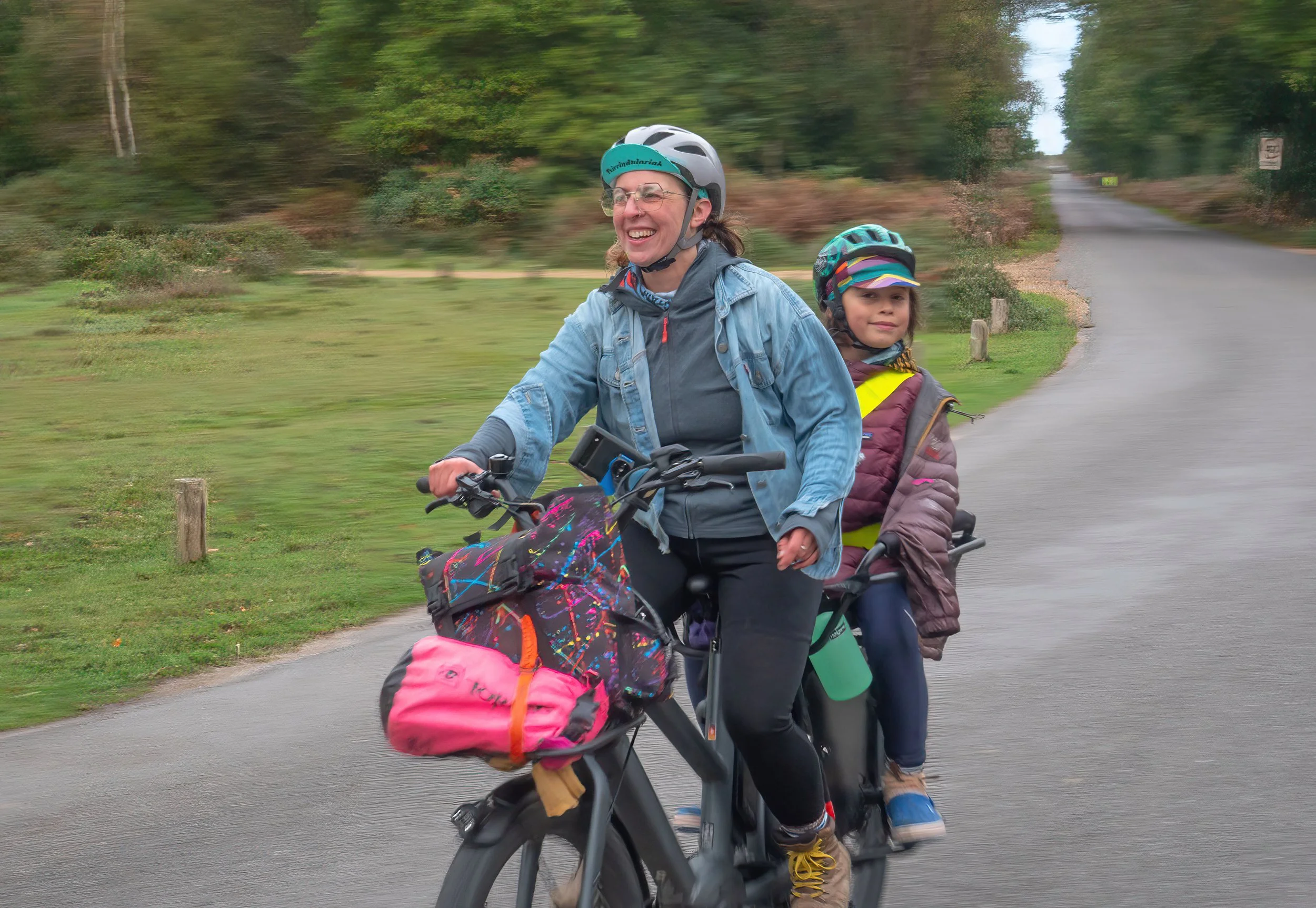 Two women riding a bicycle with a child seated in a child seat, on a paved road in a green outdoor area, all wearing helmets and smiling.