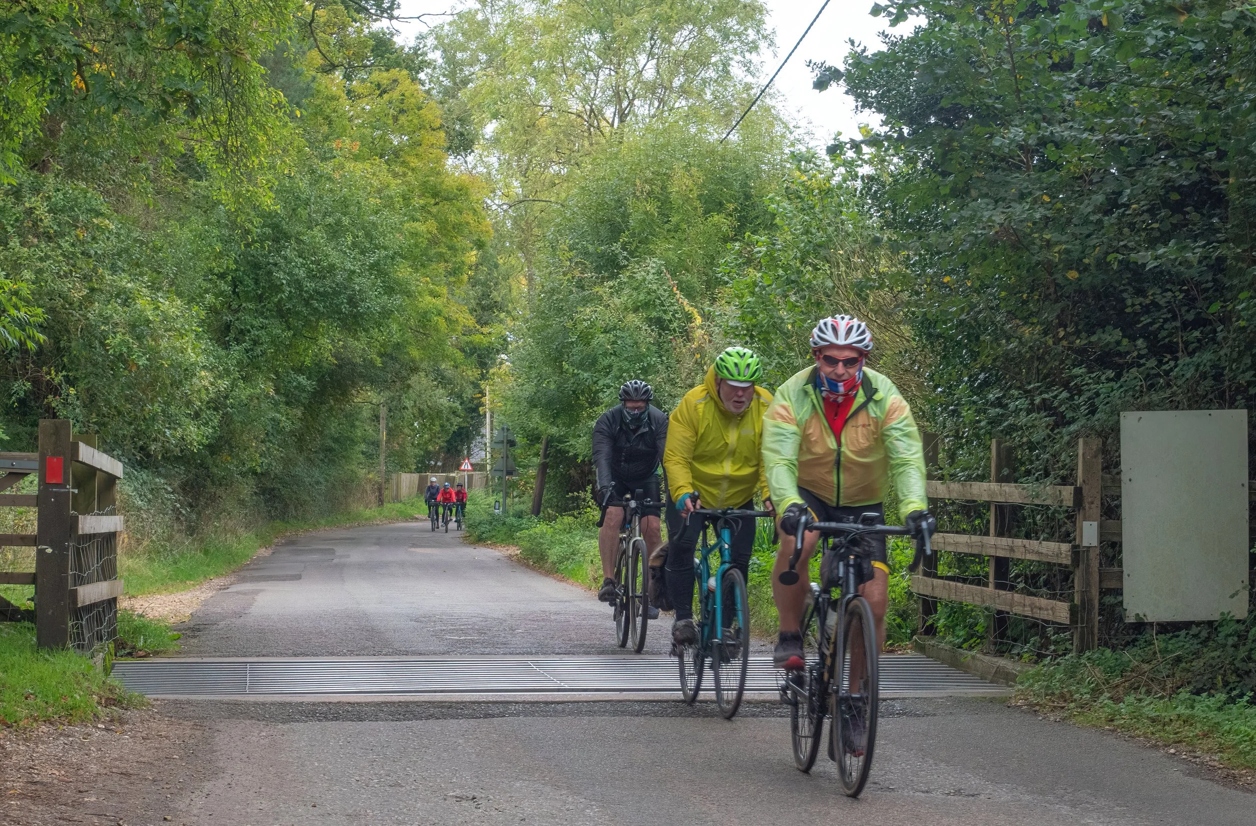 A group of five people riding bicycles on a paved trail surrounded by green trees, with four of them wearing helmets and cycling gear, and the background shows more cyclists further along the trail.