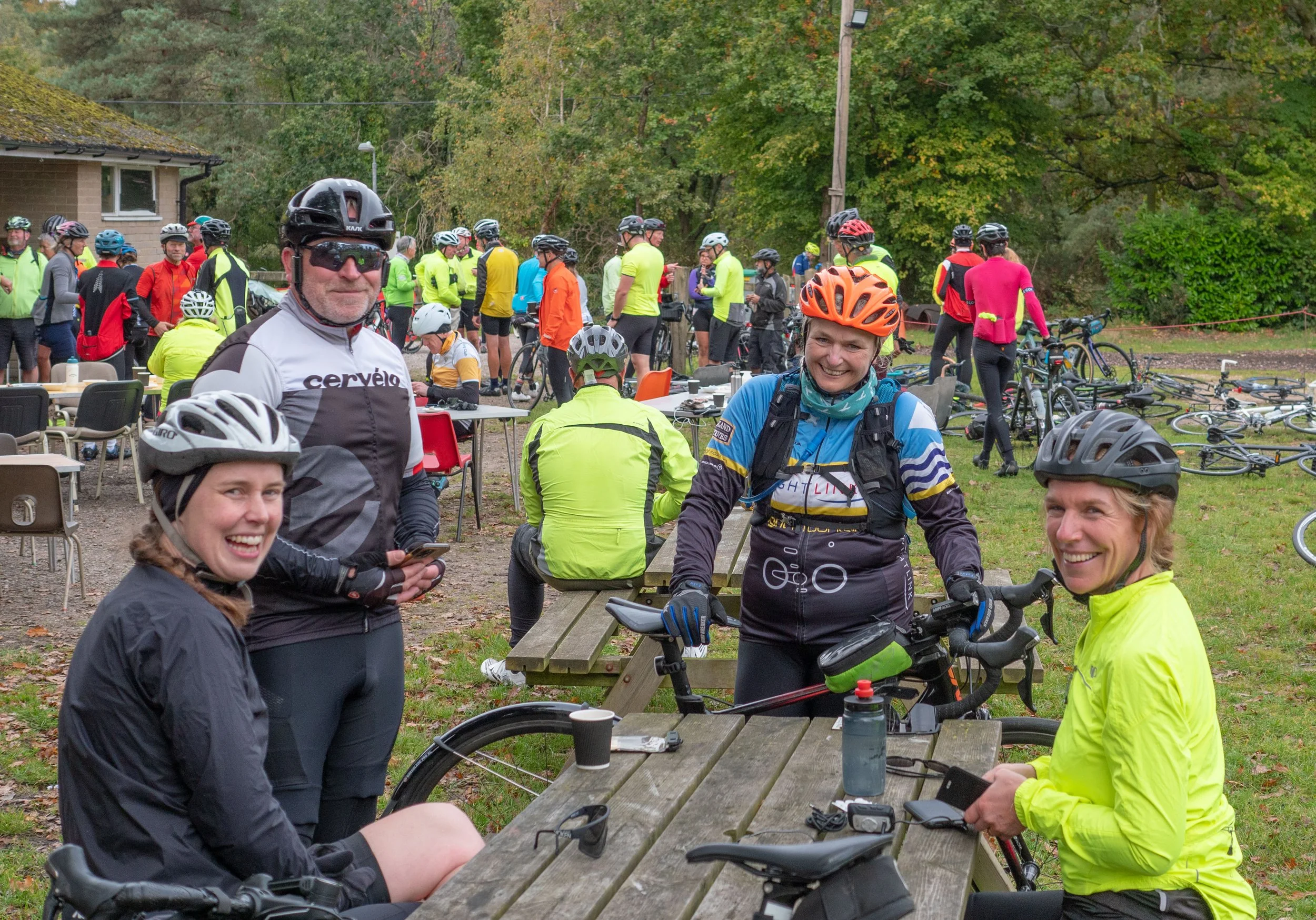 Group of cyclists gathered around outdoor tables in a park, with trees in the background.