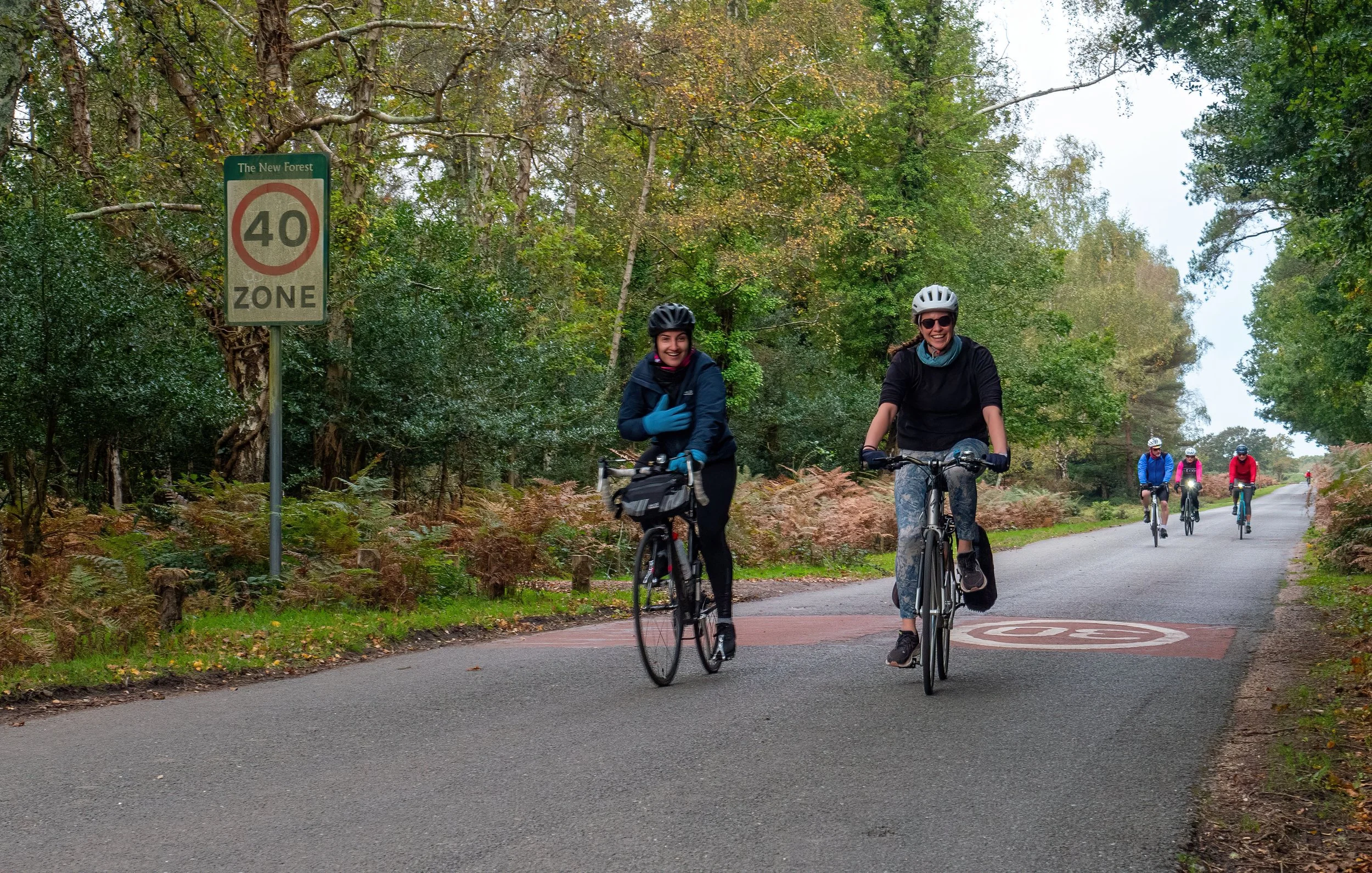 Two women riding bicycles on a forested path, smiling and wearing helmets, with a group of cyclists in the background. A sign indicating a speed zone of 40 mph is visible on the left.
