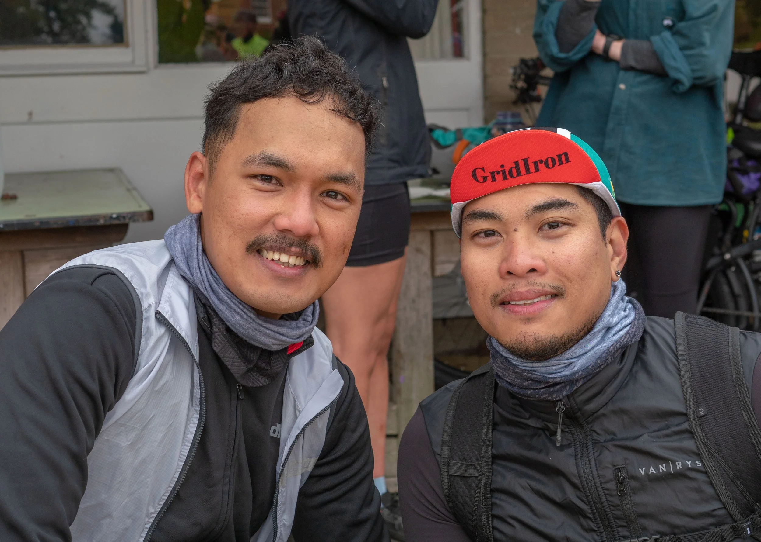 Two young men smiling at the camera, outdoors in casual winter clothing, with other people in the background.