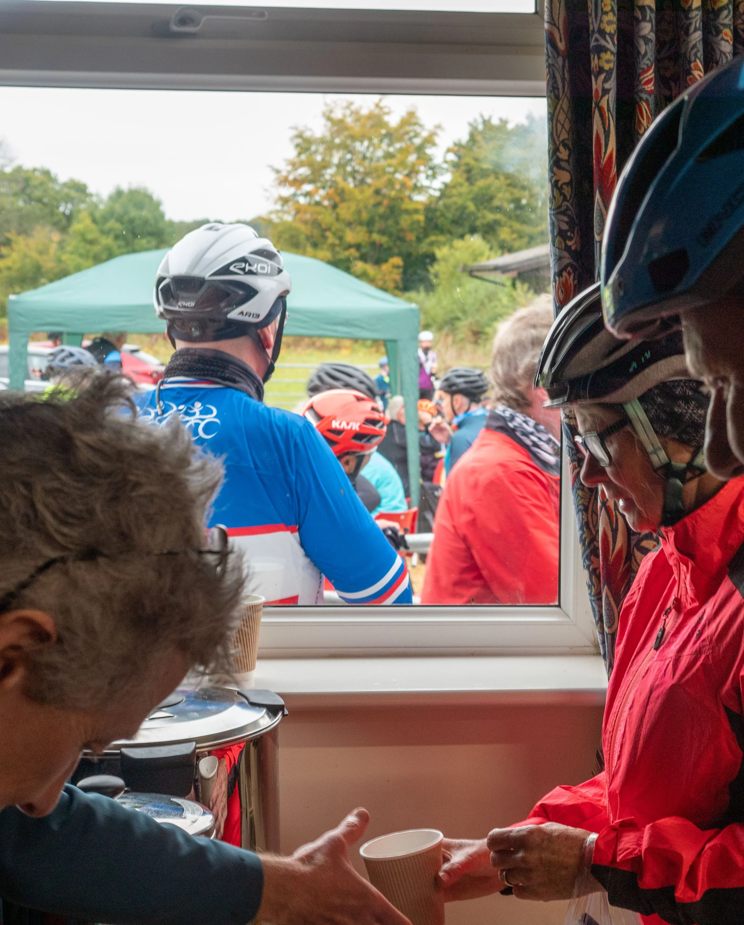 People wearing bicycle helmets inside a food truck, handing cups of coffee or drinks to each other through a window, with outdoor scenery and trees in the background.