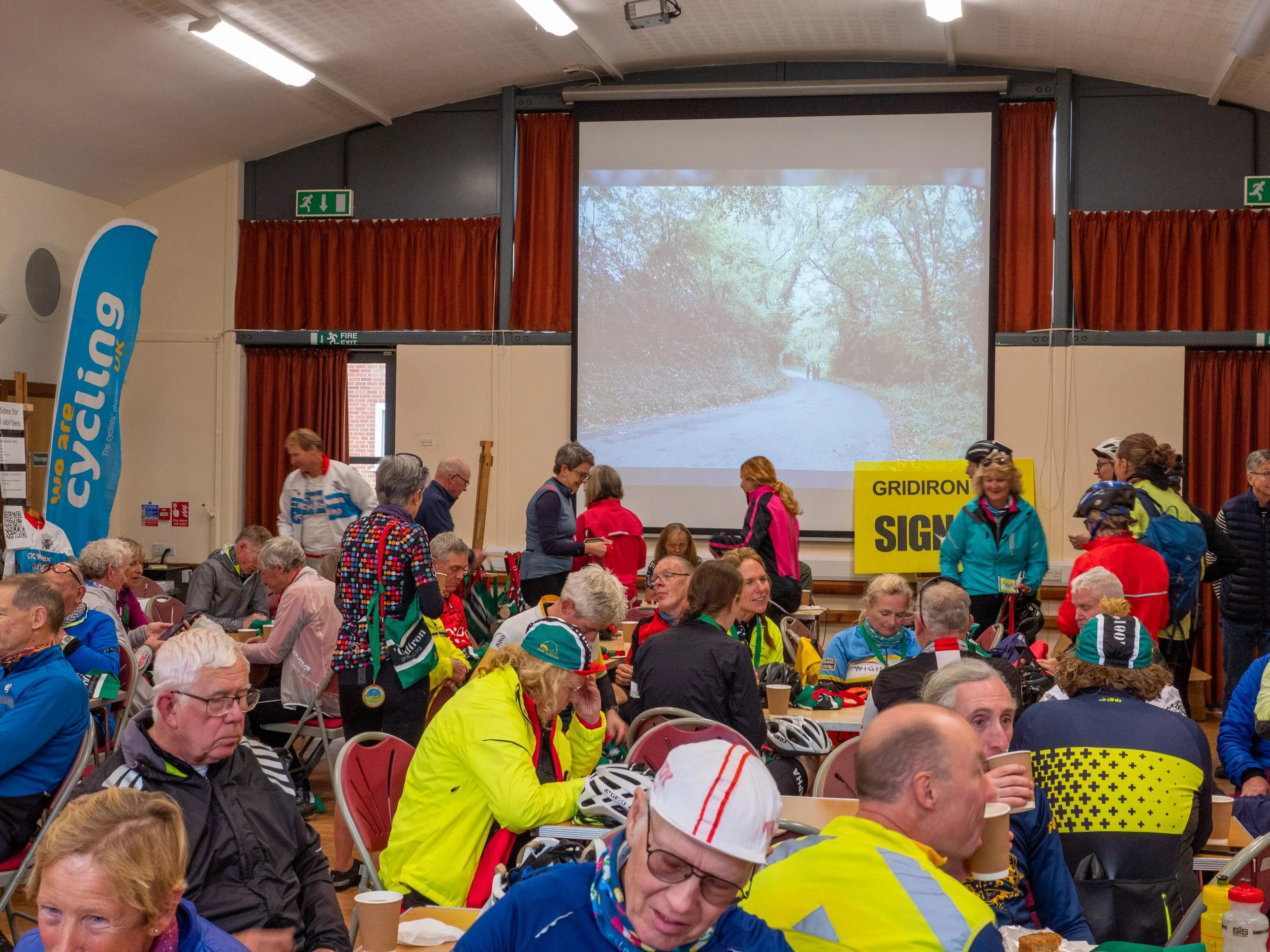 Indoor event with many people seated and standing, some wearing cycling helmets and gear. A large screen at the front shows a path through a wooded area. There are various signs and banners, including a yellow sign that says 'GRIDIRON.'