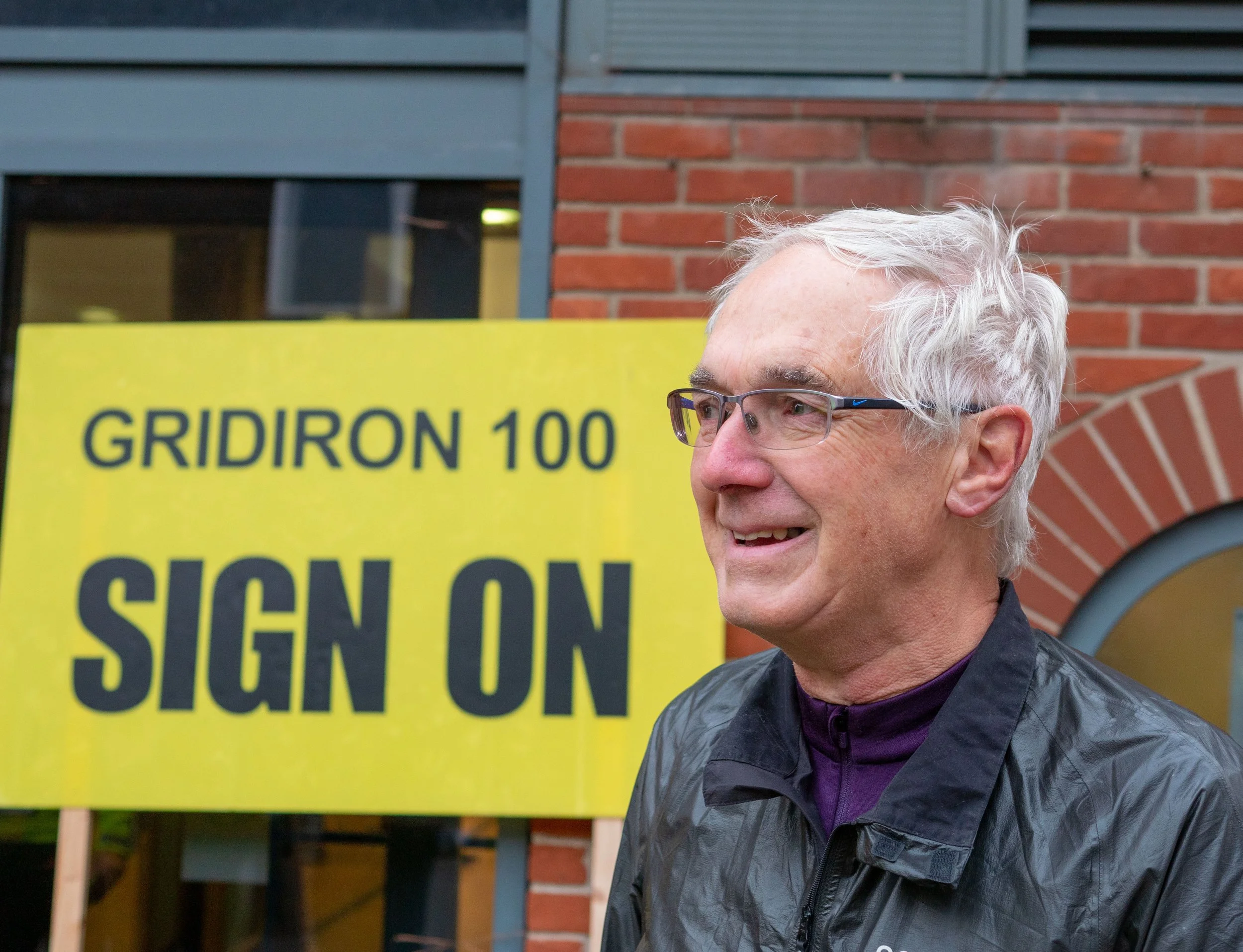 A smiling older man with glasses and white hair standing outdoors near a yellow protest sign that reads 'GRIDIRON 100 SLOW S' in front of a brick building.