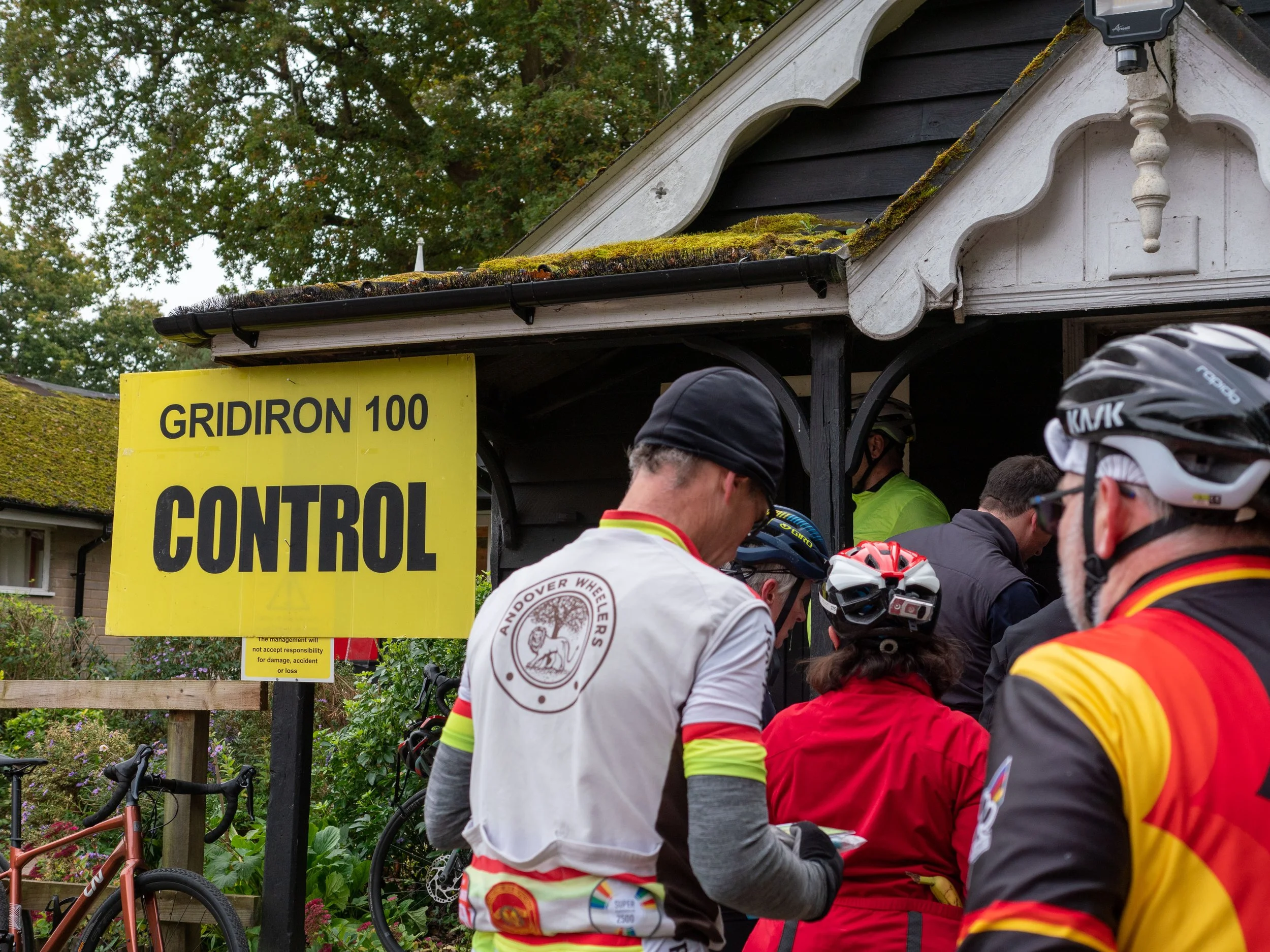 Cyclists gather under a yellow sign that reads 'GRIDIRON 100 CONTROL' with some trees and a house roof in the background.