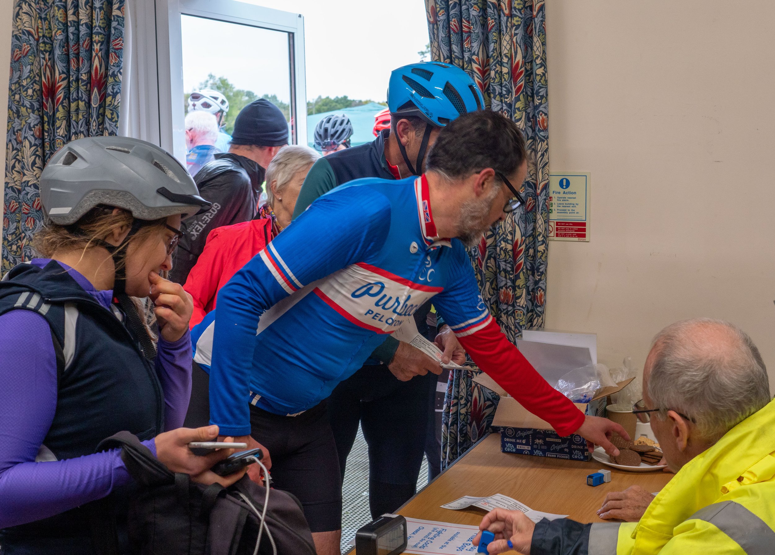 Cyclists in helmets and athletic gear gather indoors, receiving instructions or registration materials from staff at a table, with more cyclists visible outside through a window.