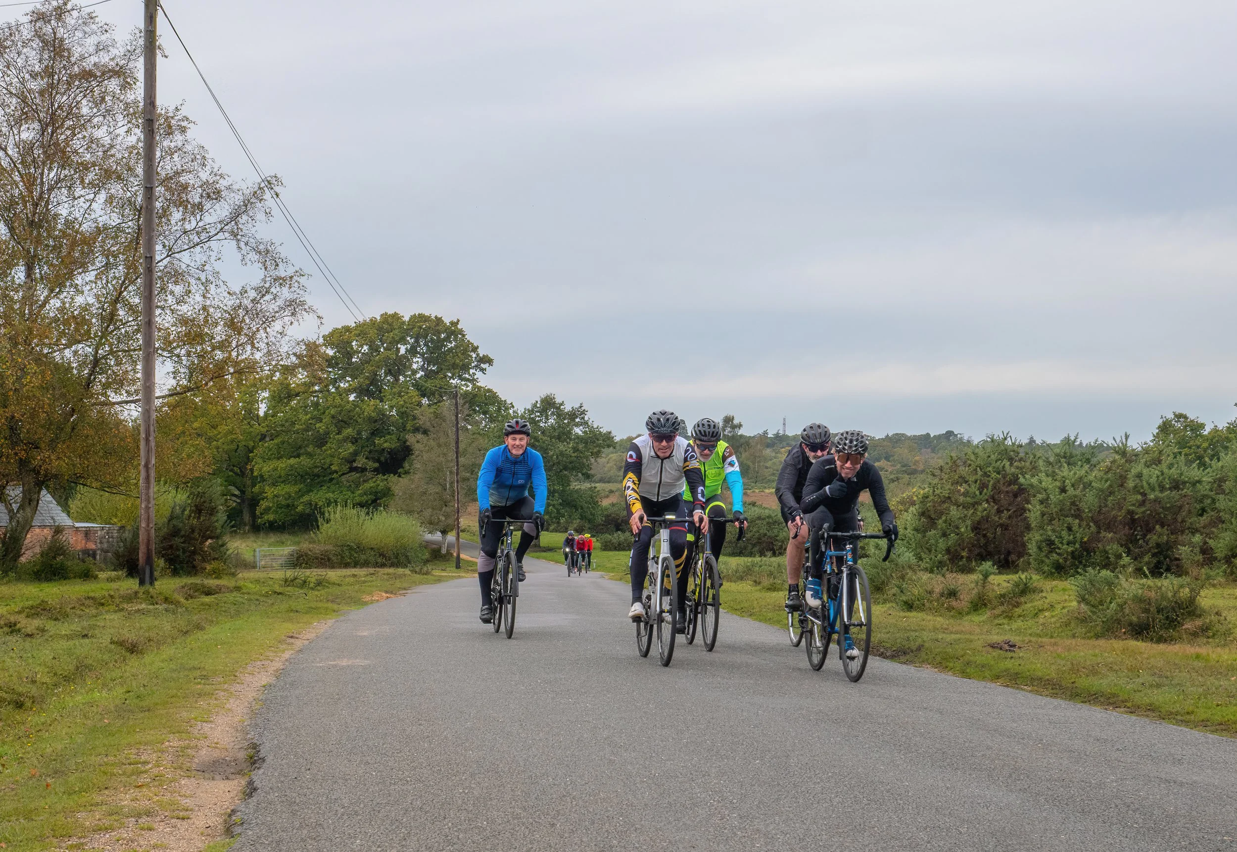 A group of five cyclists riding on a paved trail in a rural area with trees and fields, under a cloudy sky.
