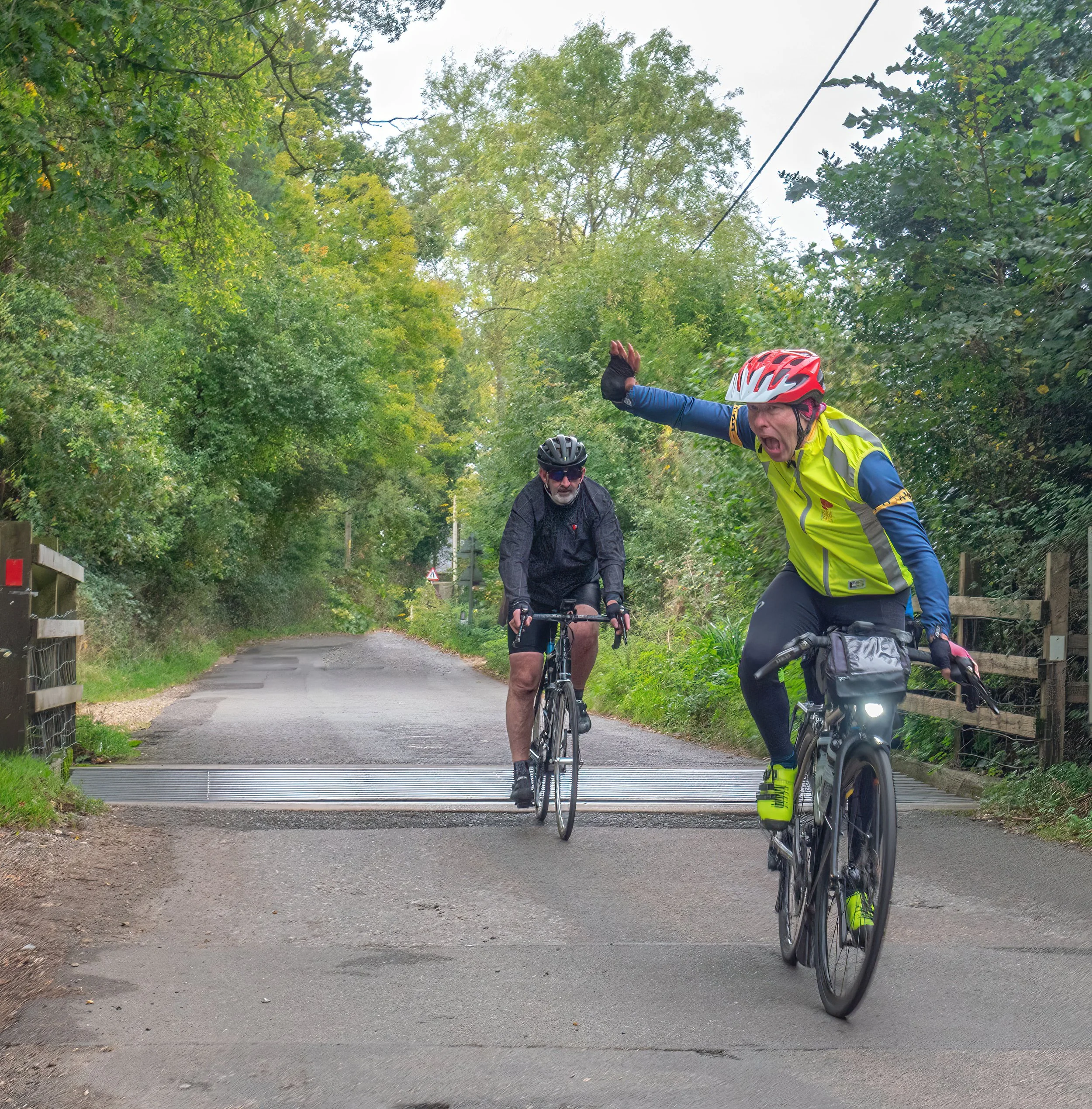 Two cyclists riding on a forested path, one wearing a yellow vest and red helmet with an expression of excitement or surprise, the other wearing a black jacket and helmet, with lush green trees on either side.