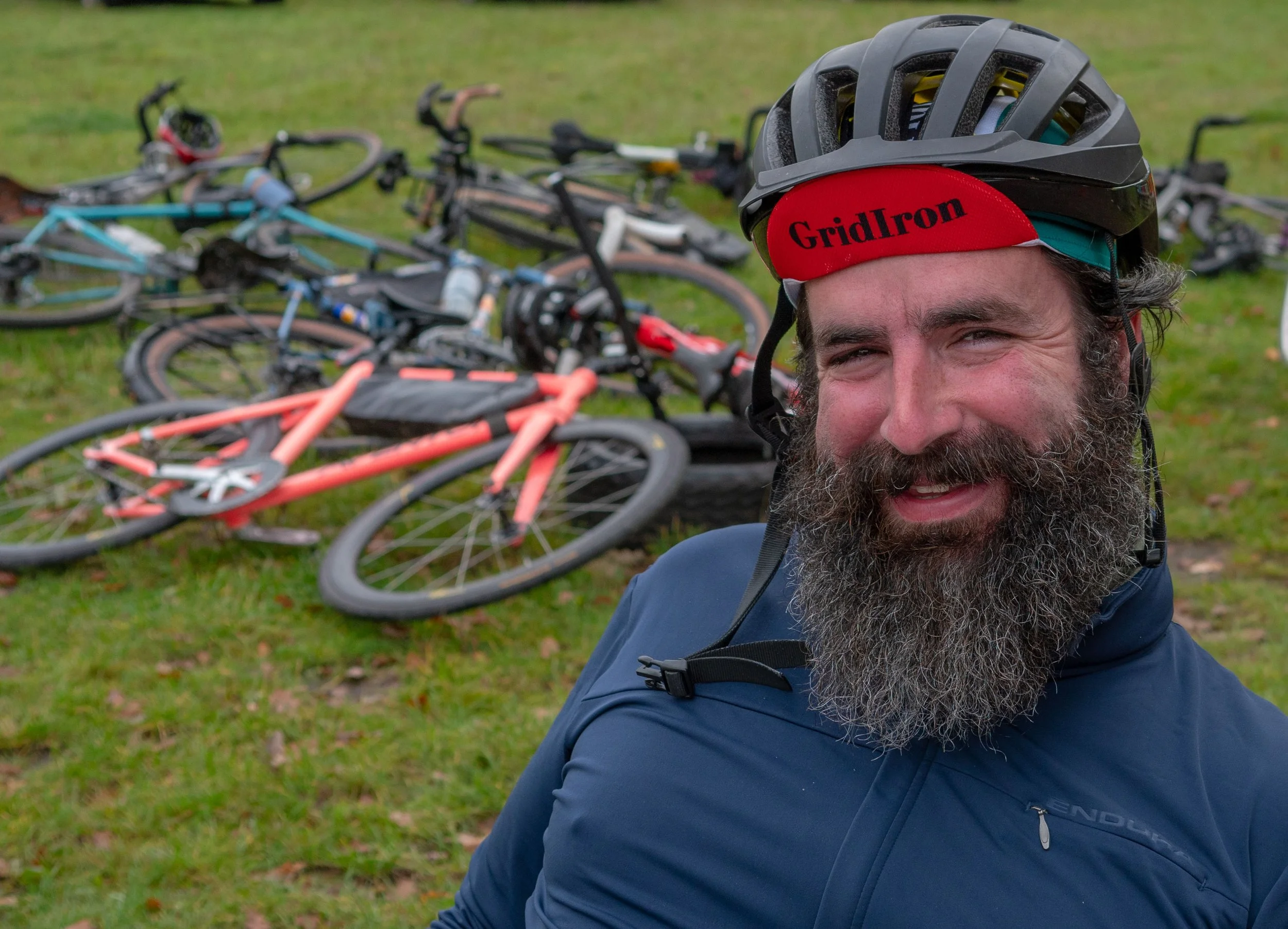 Close-up of a smiling man wearing a black helmet, red headband with the word 'GridIron', and a blue jacket, with bicycles lying on grass in the background.