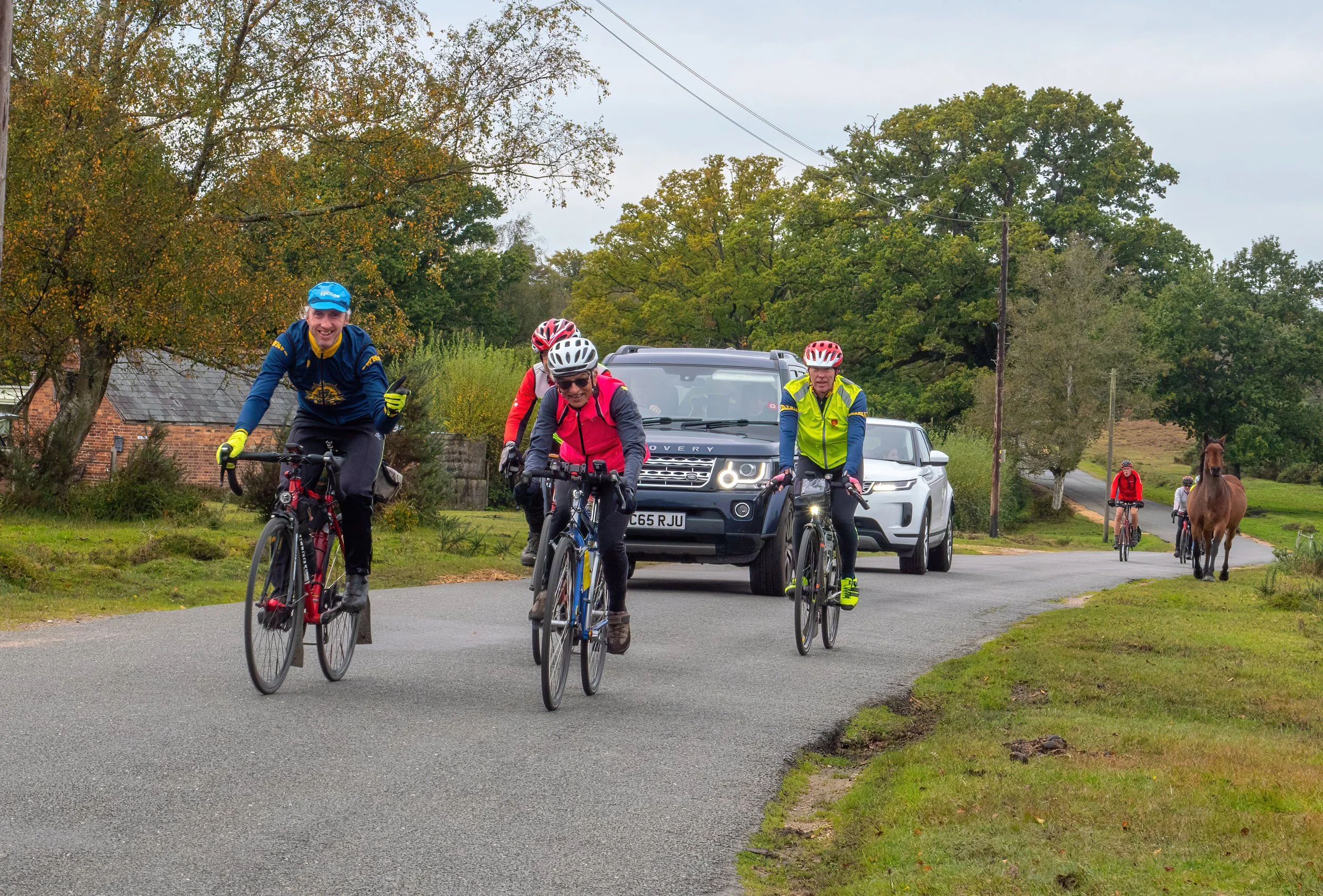 Group of people riding bicycles on a rural road, with cars and horses in the background, surrounded by trees and overcast sky.