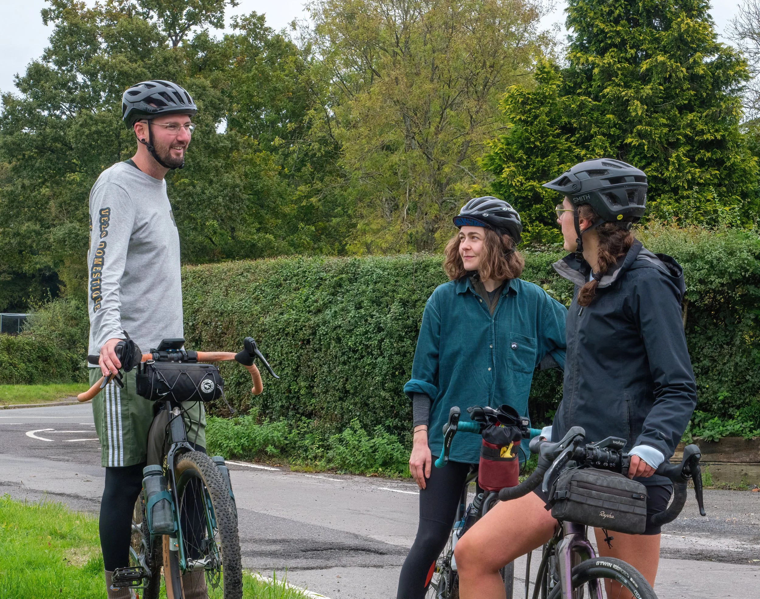Three cyclists, two women and one man, wearing helmets and talking outdoors in a park with green trees and grass, with bikes.