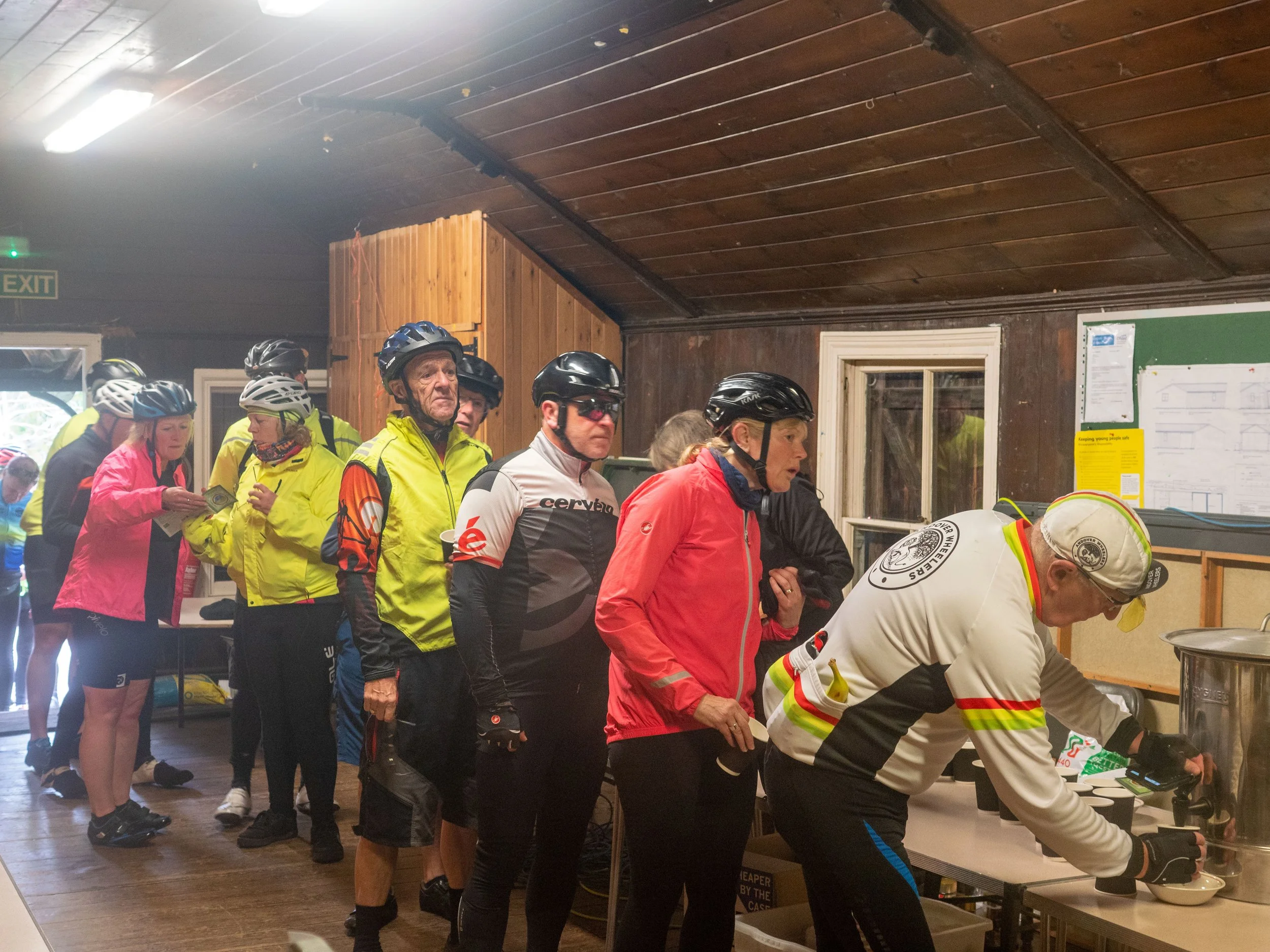 A group of people wearing cycling gear and helmets standing in line at a table inside a wooden-ceiling room, possibly for food or registration.