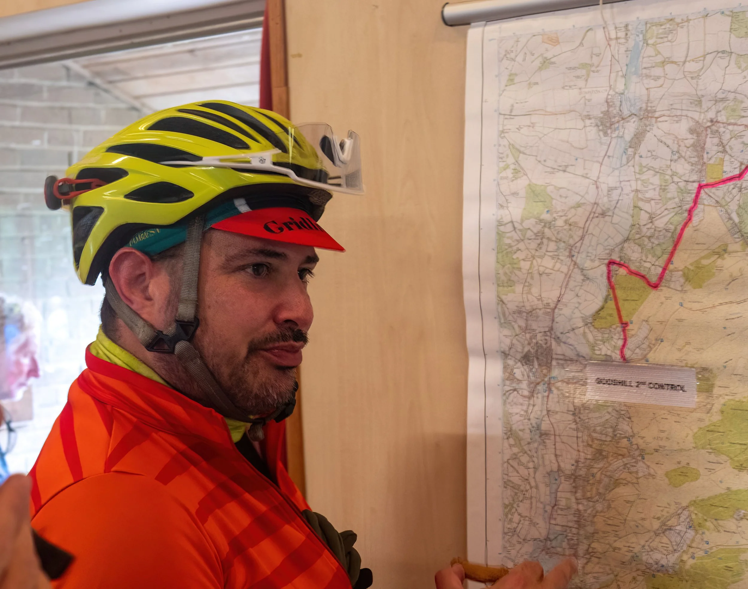 A man wearing a yellow and black bicycle helmet and an orange jacket is looking at a large map on a wall, with a focus on the map and his profile visible.