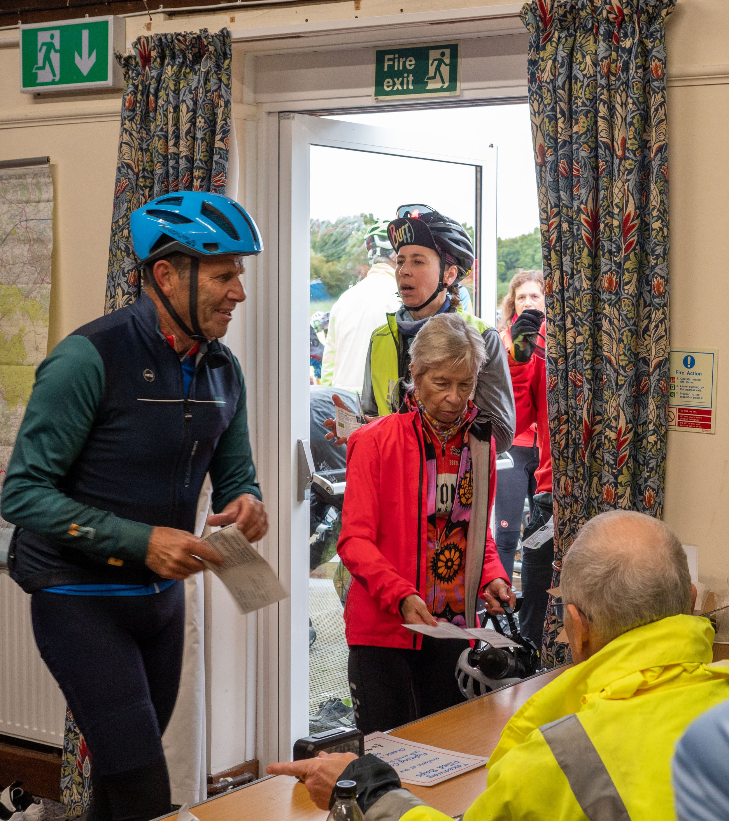 People wearing cycling helmets and jackets inside a building near a fire exit, with one person reading a paper and others engaged in conversation.