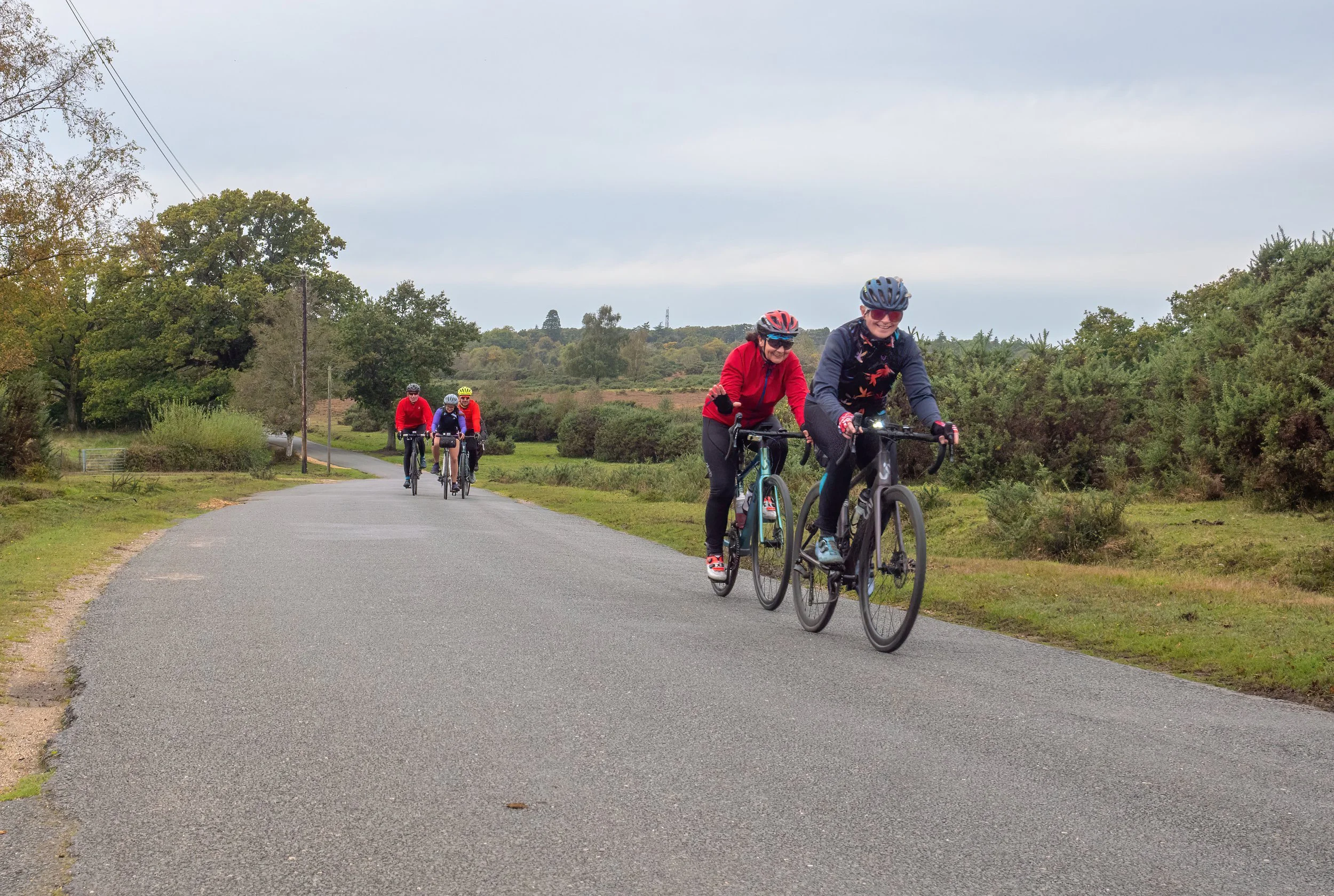 Four people riding bicycles on a rural road, wearing helmets and colorful jackets, with lush green trees and cloudy sky in the background.