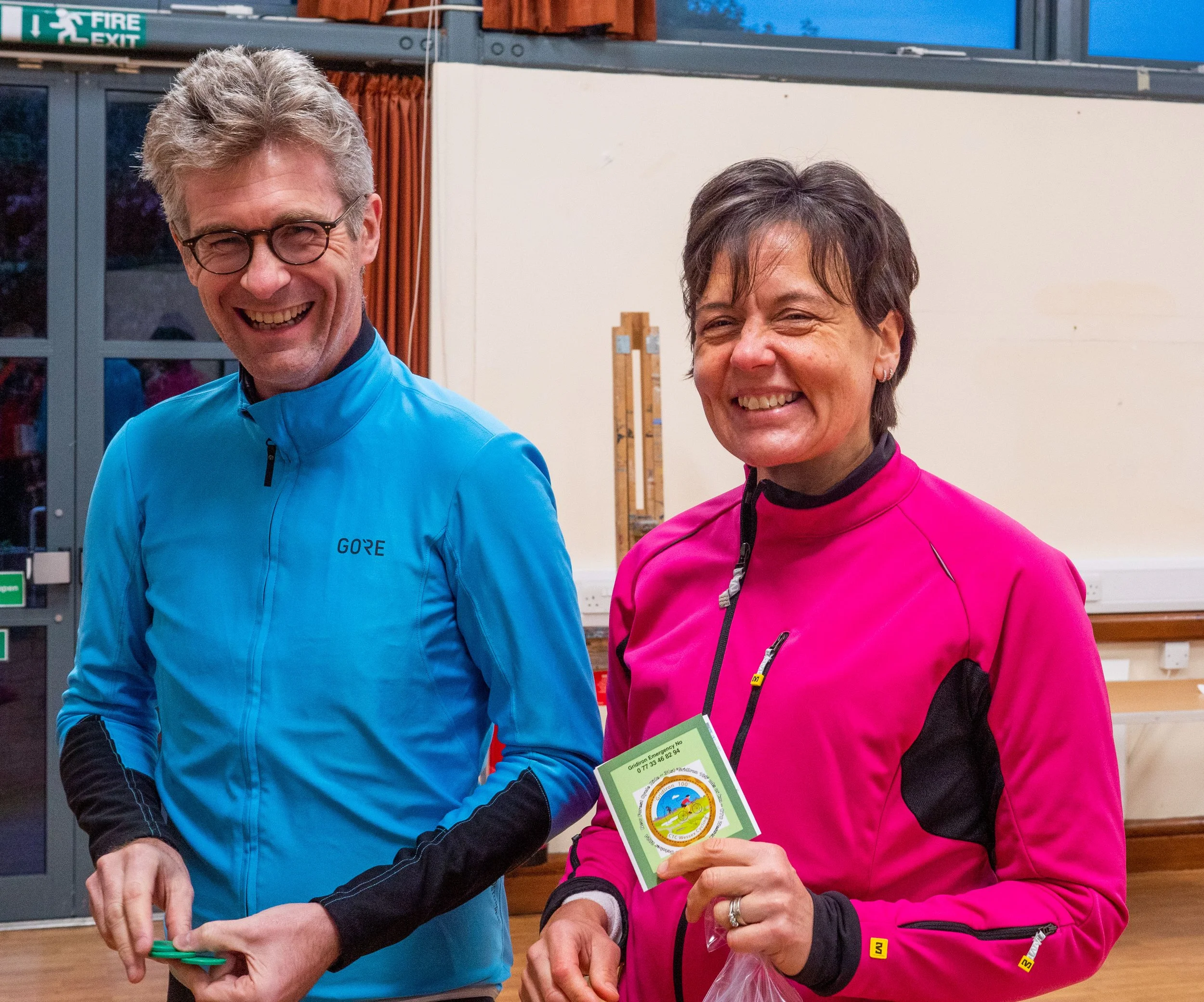 Two people smiling, one wearing a blue jacket and glasses, the other in a pink jacket, holding a green and white award or certificate, indoors with a window and a green emergency exit sign in the background.