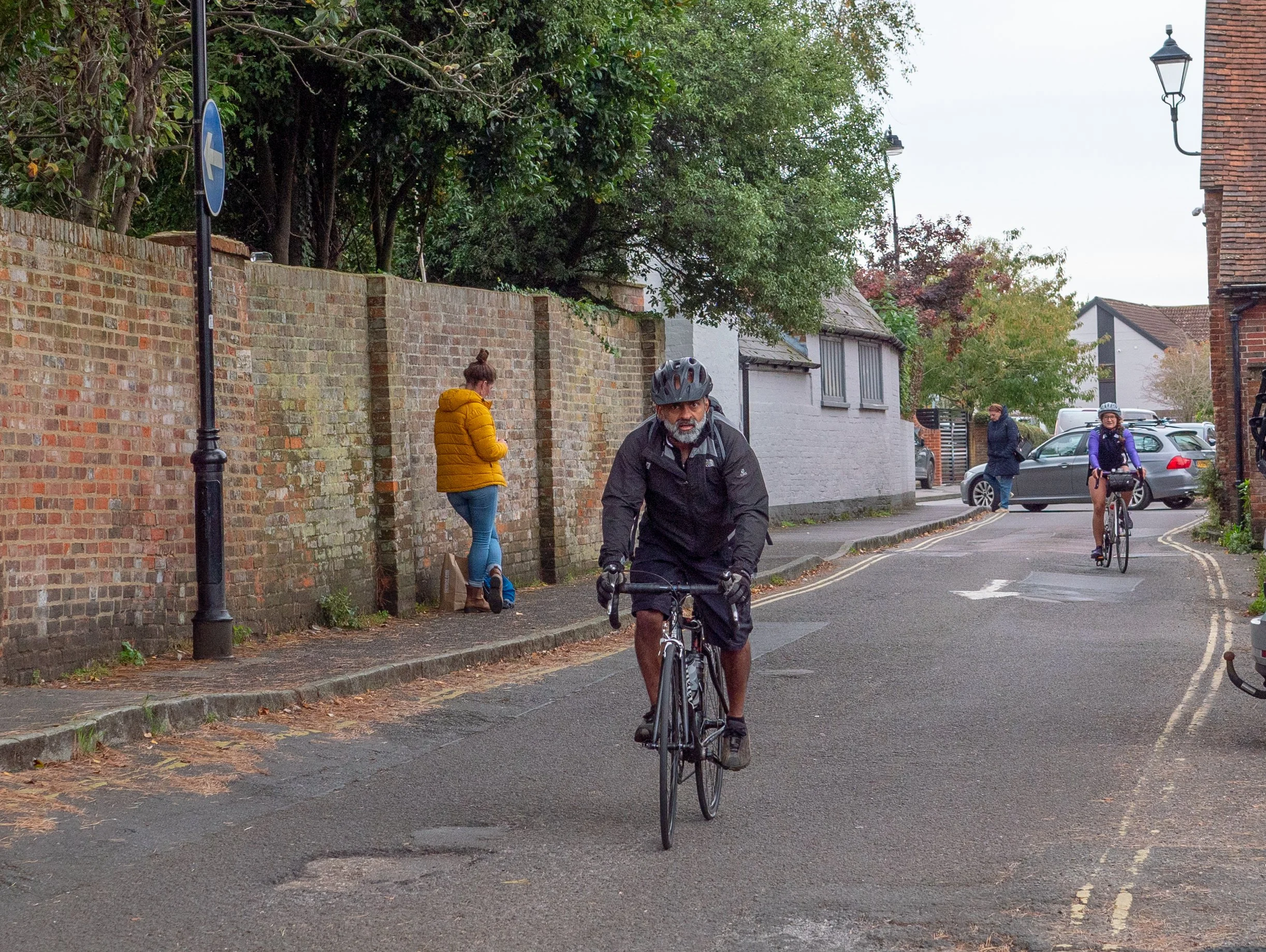 A man riding a bicycle on a city street, with three women standing and walking along the brick sidewalk and parked cars visible in the background.