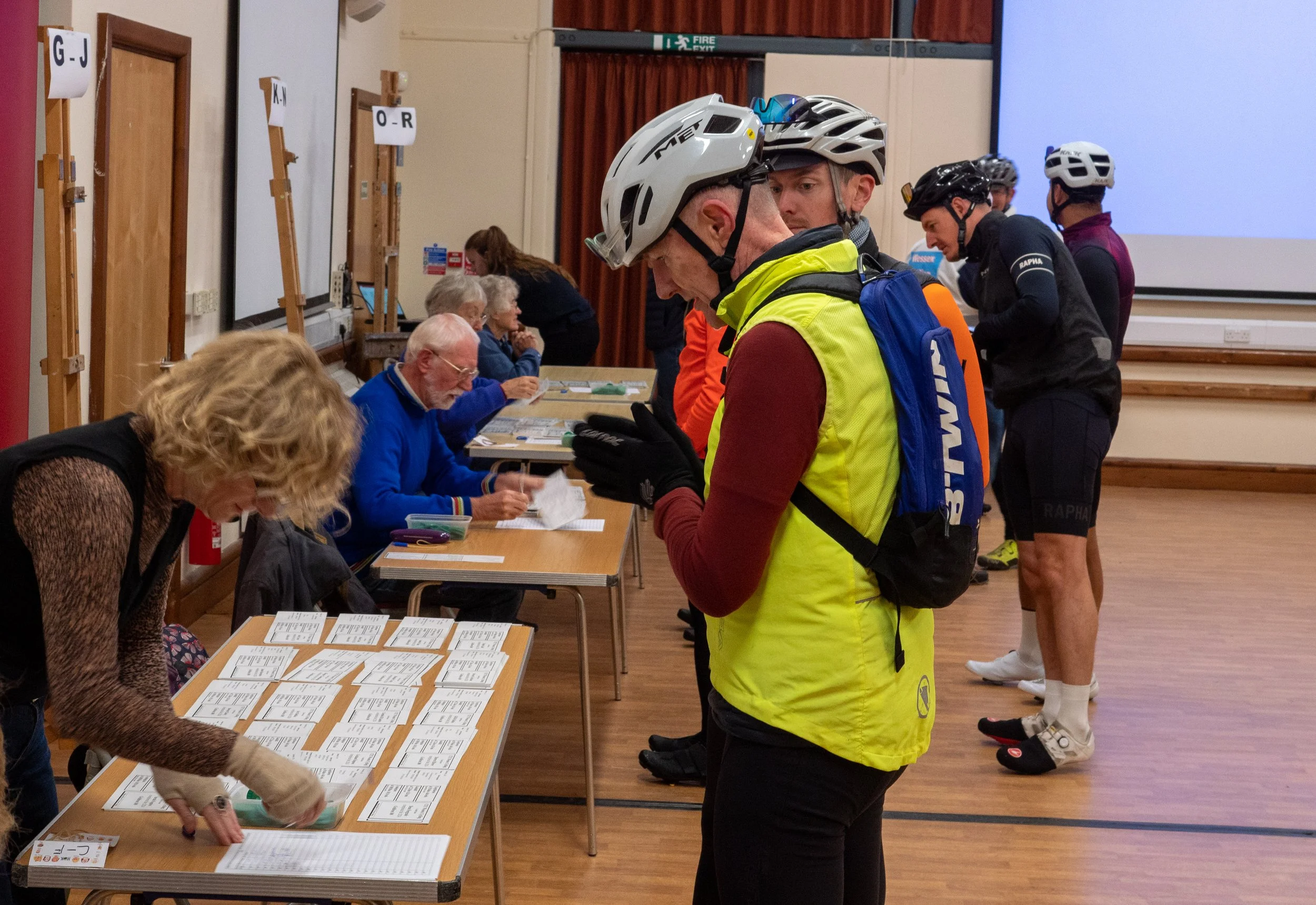 People in cycling gear registering at event tables with paperwork in a large indoor room.
