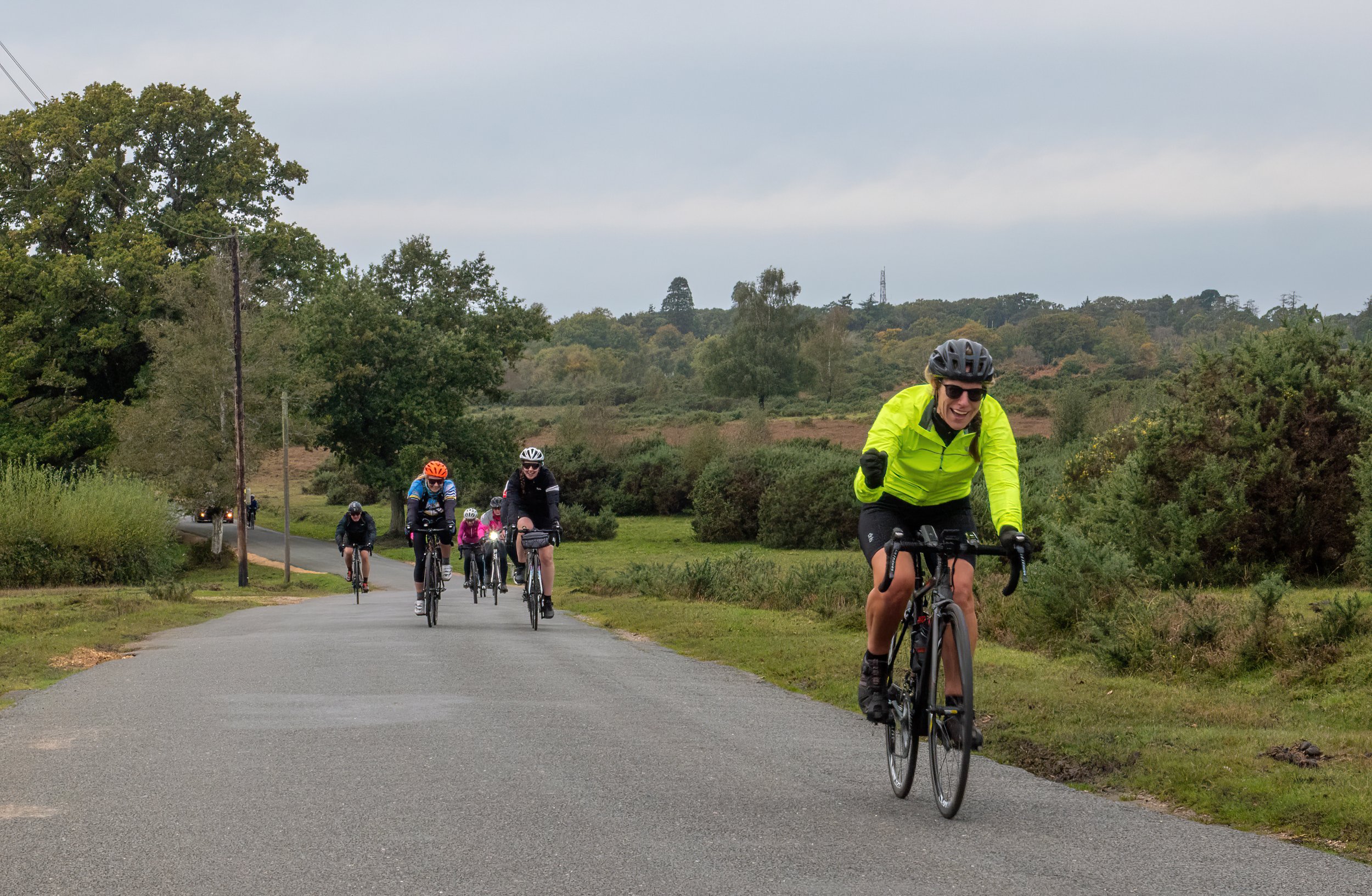 Group of cyclists riding on a paved trail through a green landscape with trees and shrubs, overcast sky.
