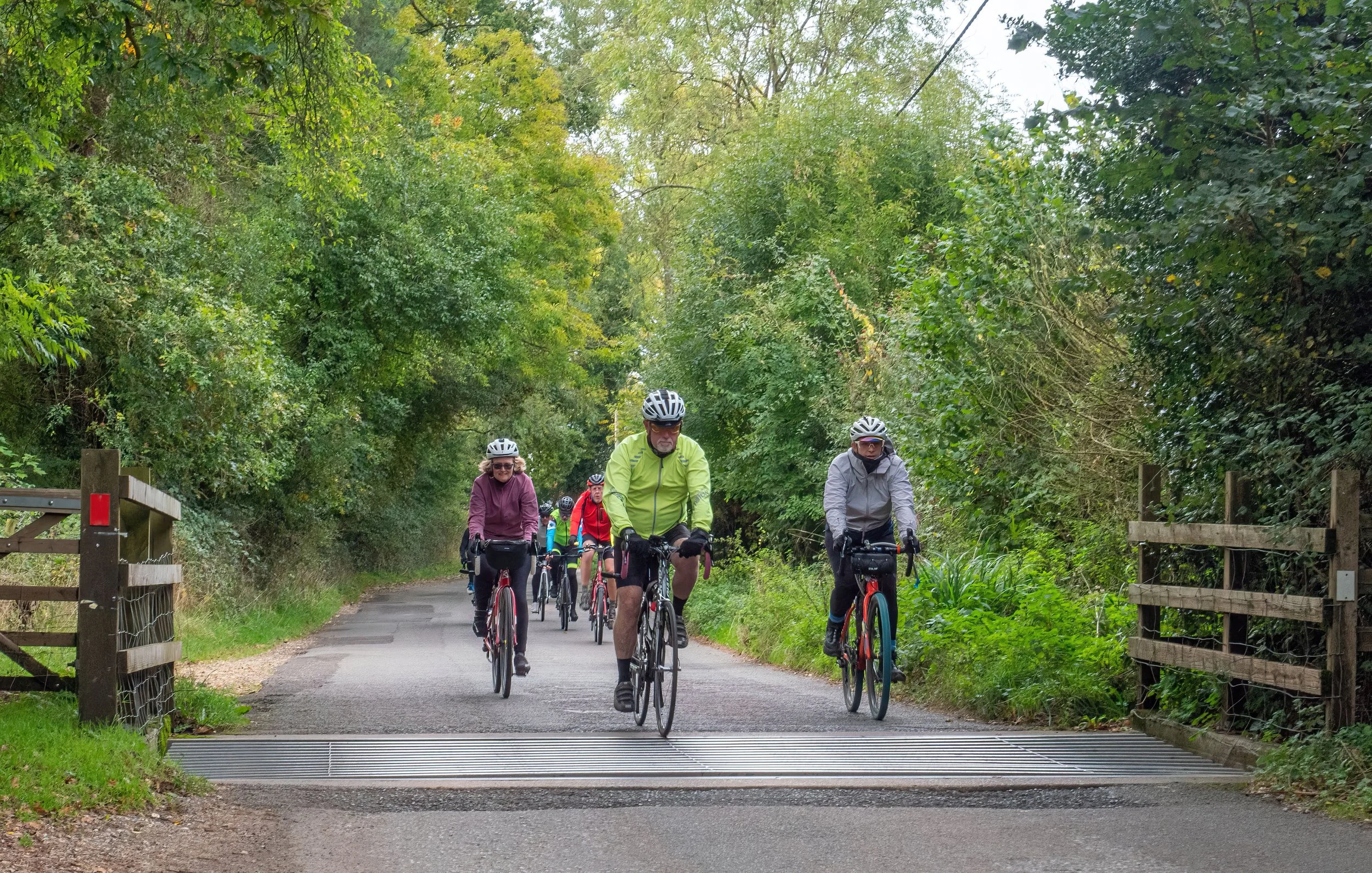 Group of people riding bicycles along a wooded path, approaching a railroad crossing with a barrier arm.