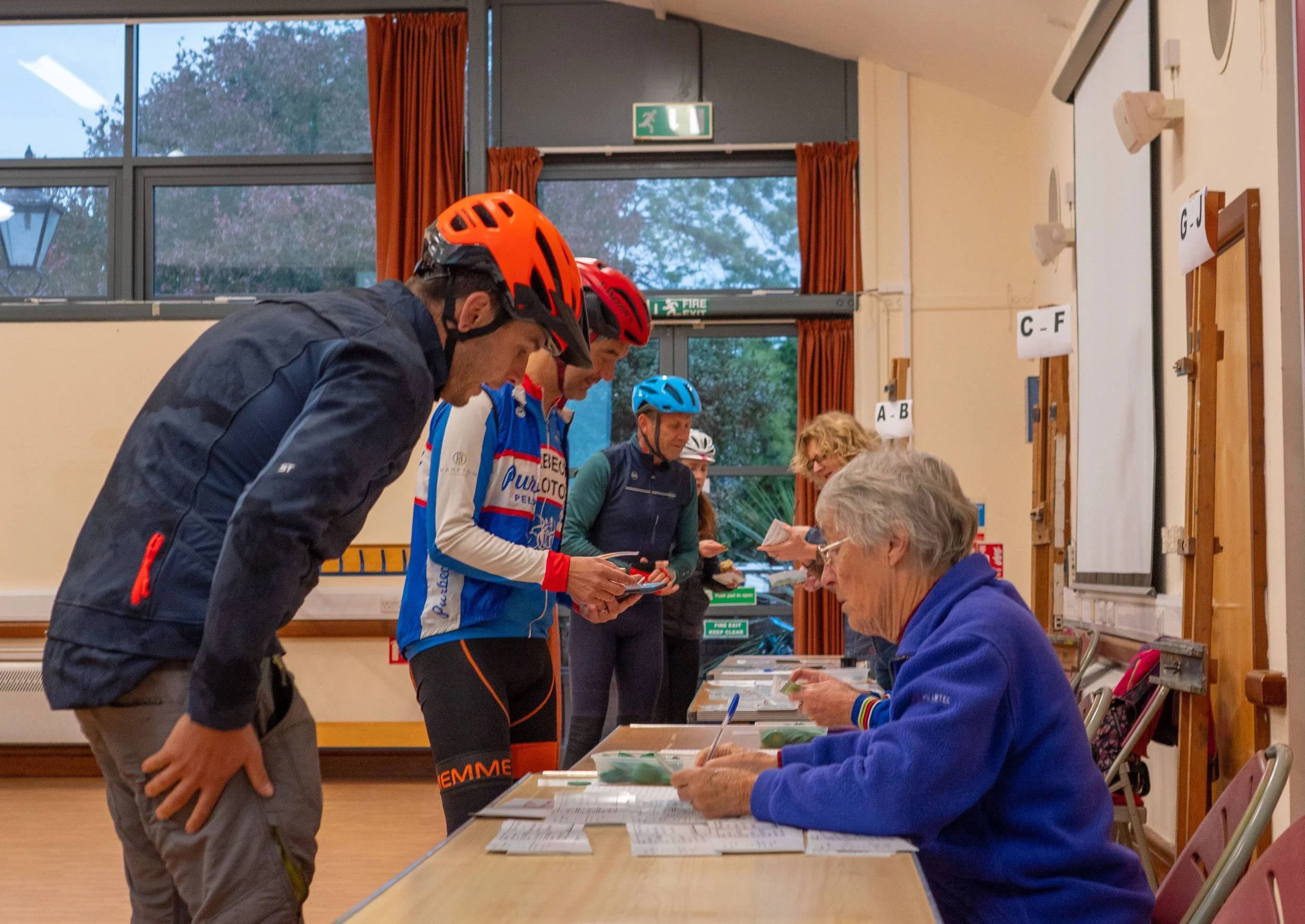 Several people wearing cycling helmets are standing in line at a registration or check-in table inside a building with large windows and orange curtains. An older woman seated at the table is assisting them with paperwork or registration material.