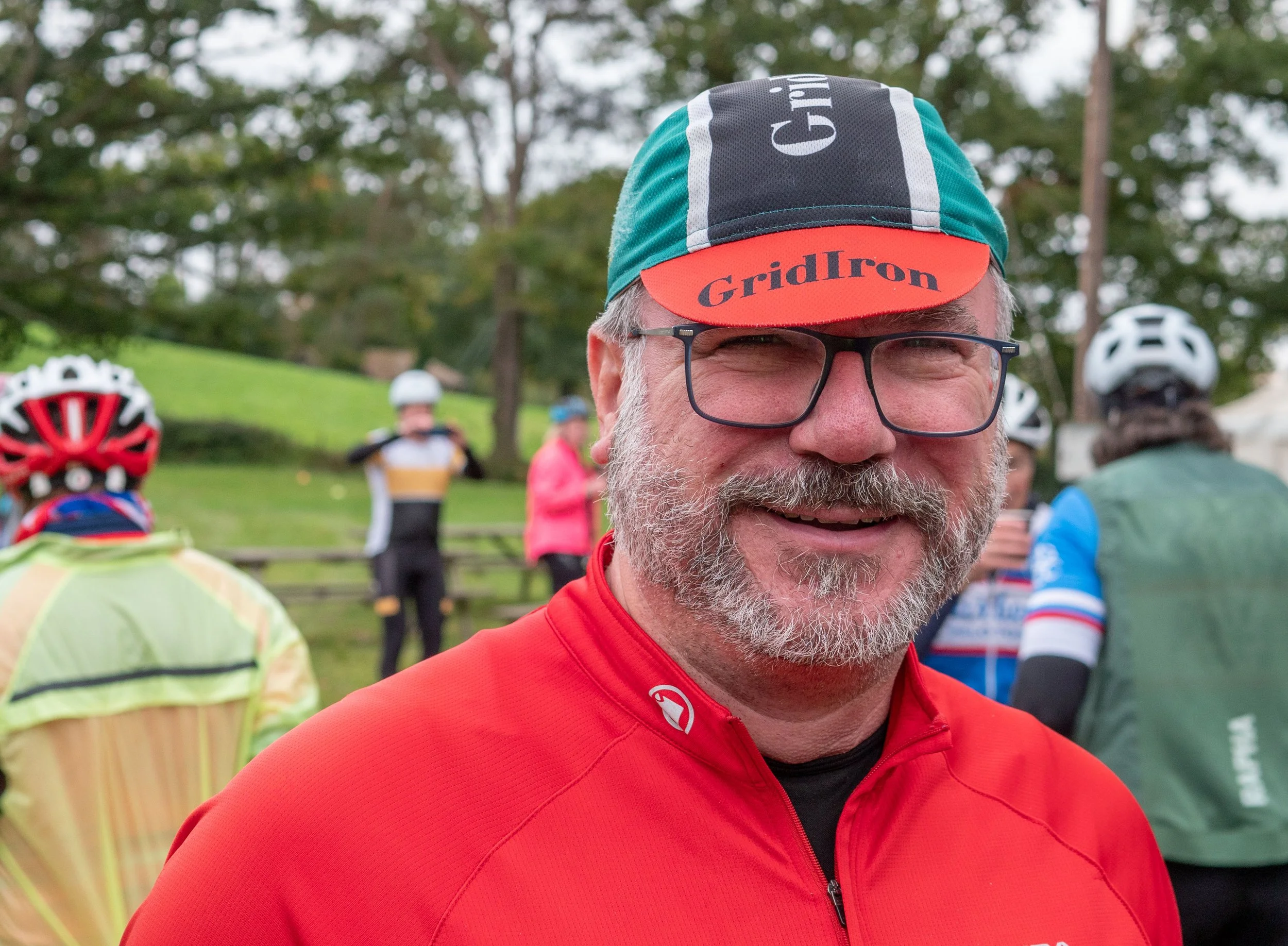 A close-up of a smiling man wearing glasses, a colorful cycling cap with the text 'GridIron', and a red cycling jacket, with blurred people and trees in the background.