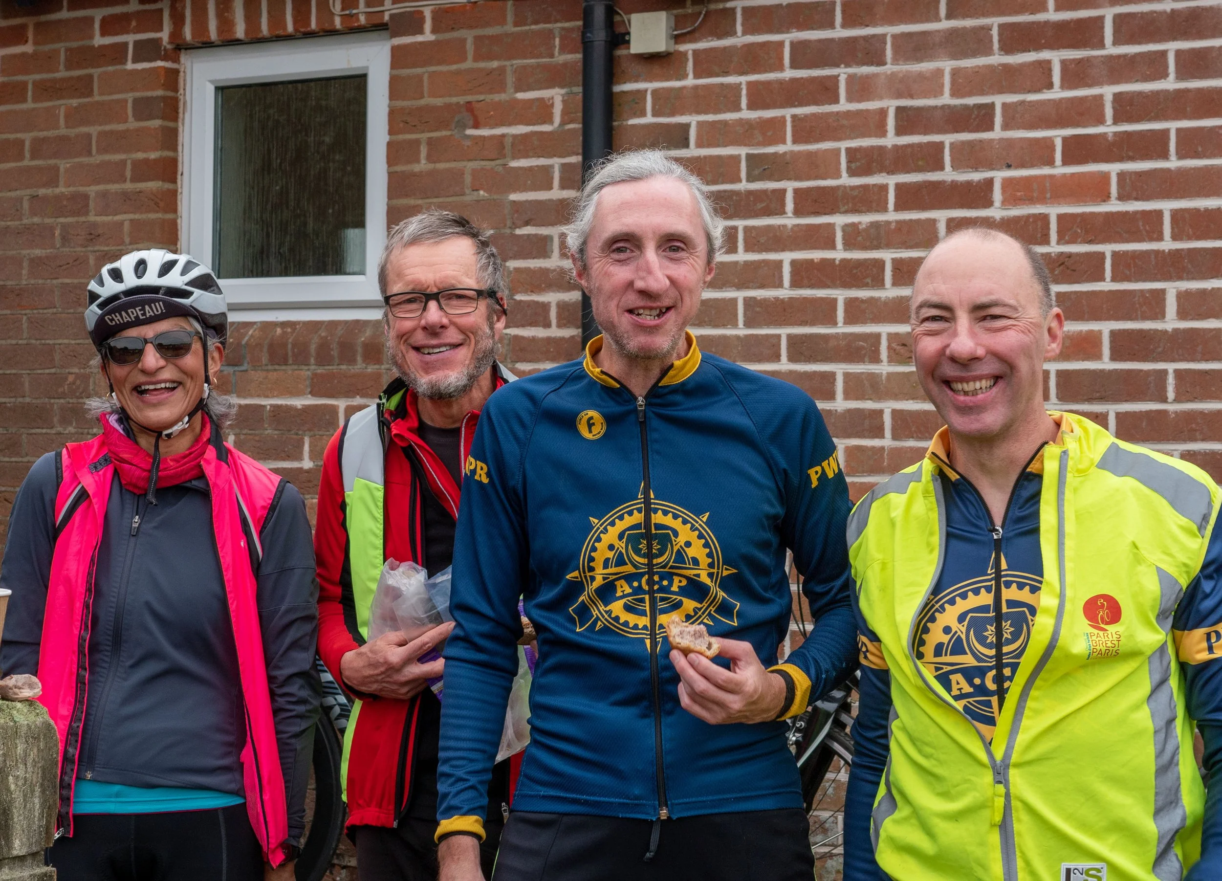 Four smiling individuals outdoors standing against a brick wall, wearing athletic clothing and gear, including helmets and reflective jackets, suggesting they are part of a cycling or running group.