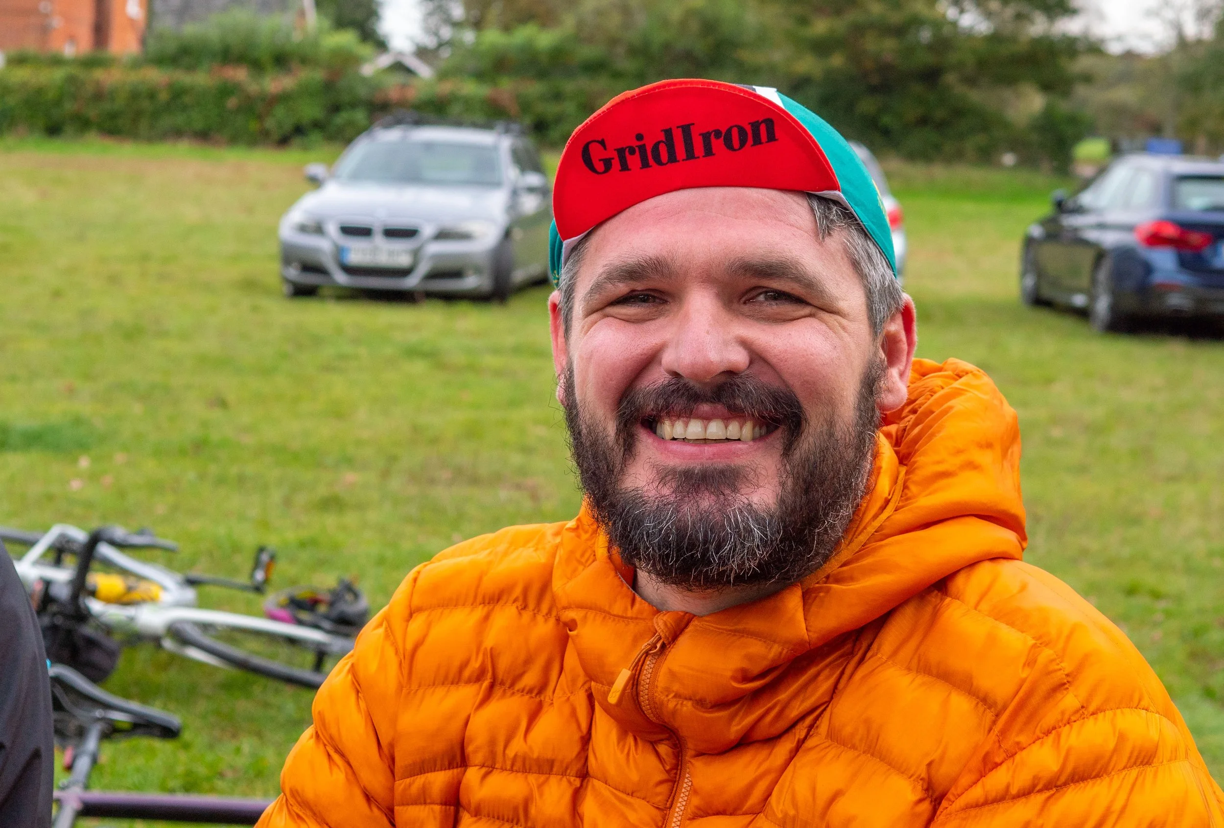 A man smiling outdoors, wearing an orange jacket and a colorful cycling cap with the word "GridIron" on it; there are bicycles and parked cars in the background.