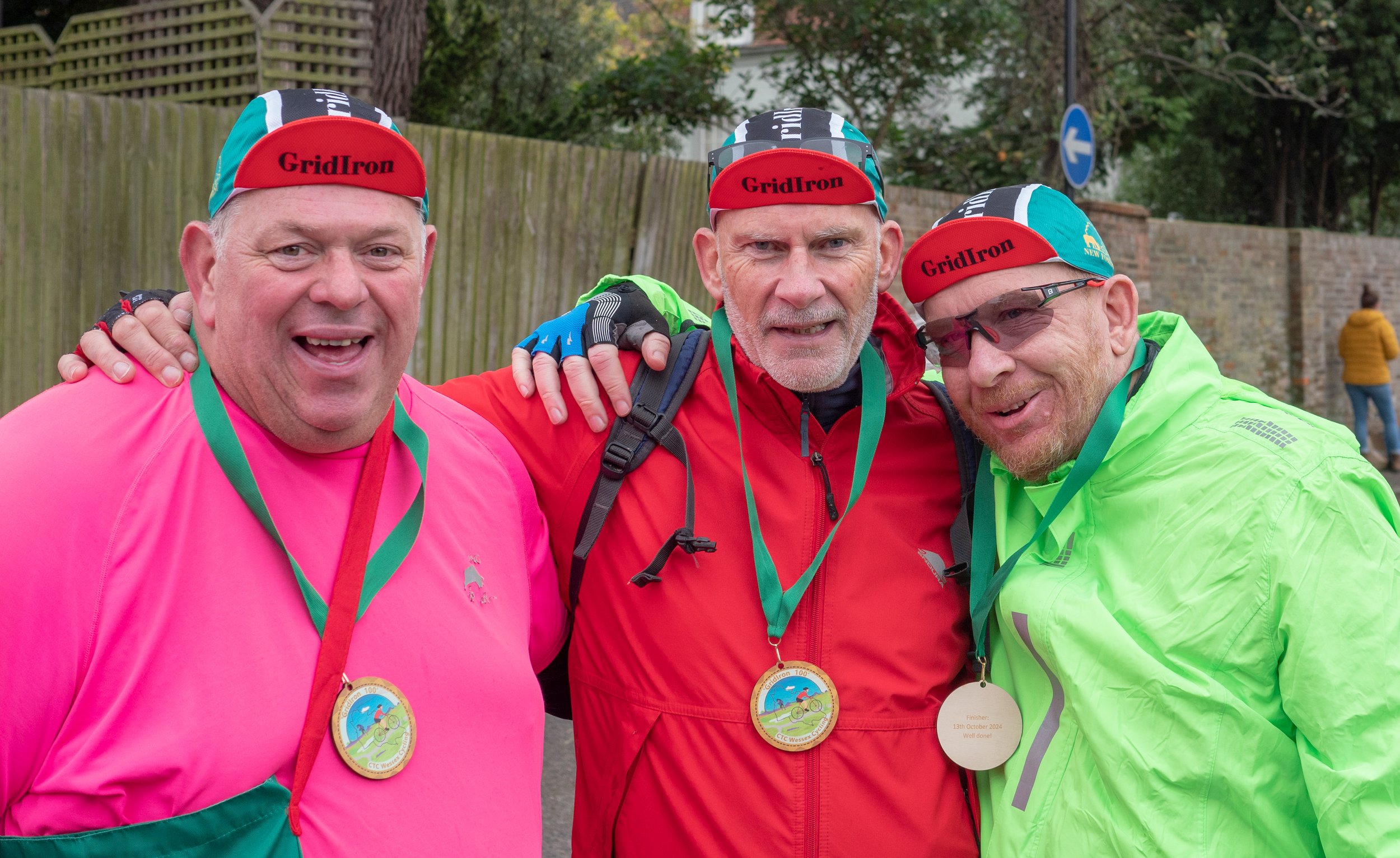 Three men wearing colorful athletic clothing and medals, smiling and posing outdoors with a wooden fence and trees in the background.
