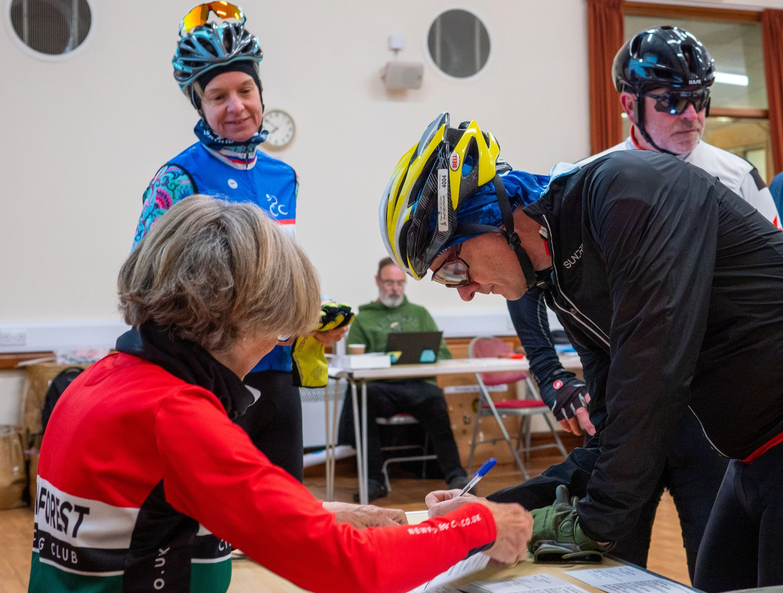 Cyclists signing papers at an indoor event, wearing helmets and cycling gear.