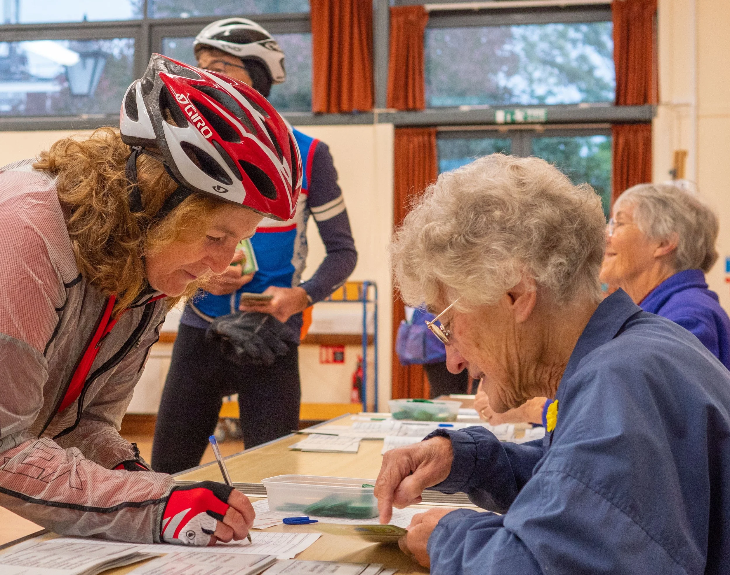 Women and elderly people seated at tables, engaging in activities, with a woman wearing a red and white bike helmet leaning over the table, in a room with large windows and orange curtains.