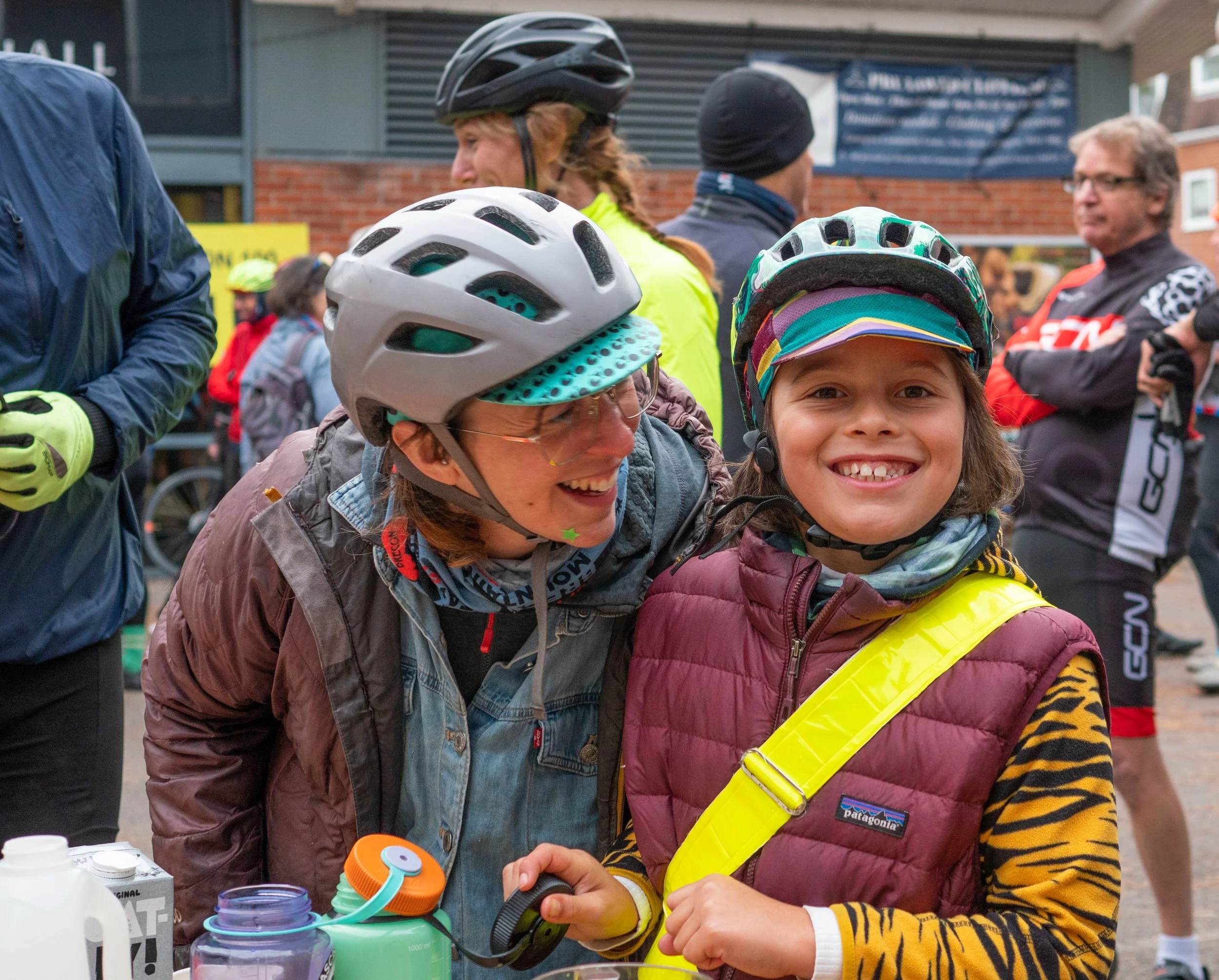 Smiling young girl and woman wearing helmets in a crowded outdoor event, with other people and a building in the background.