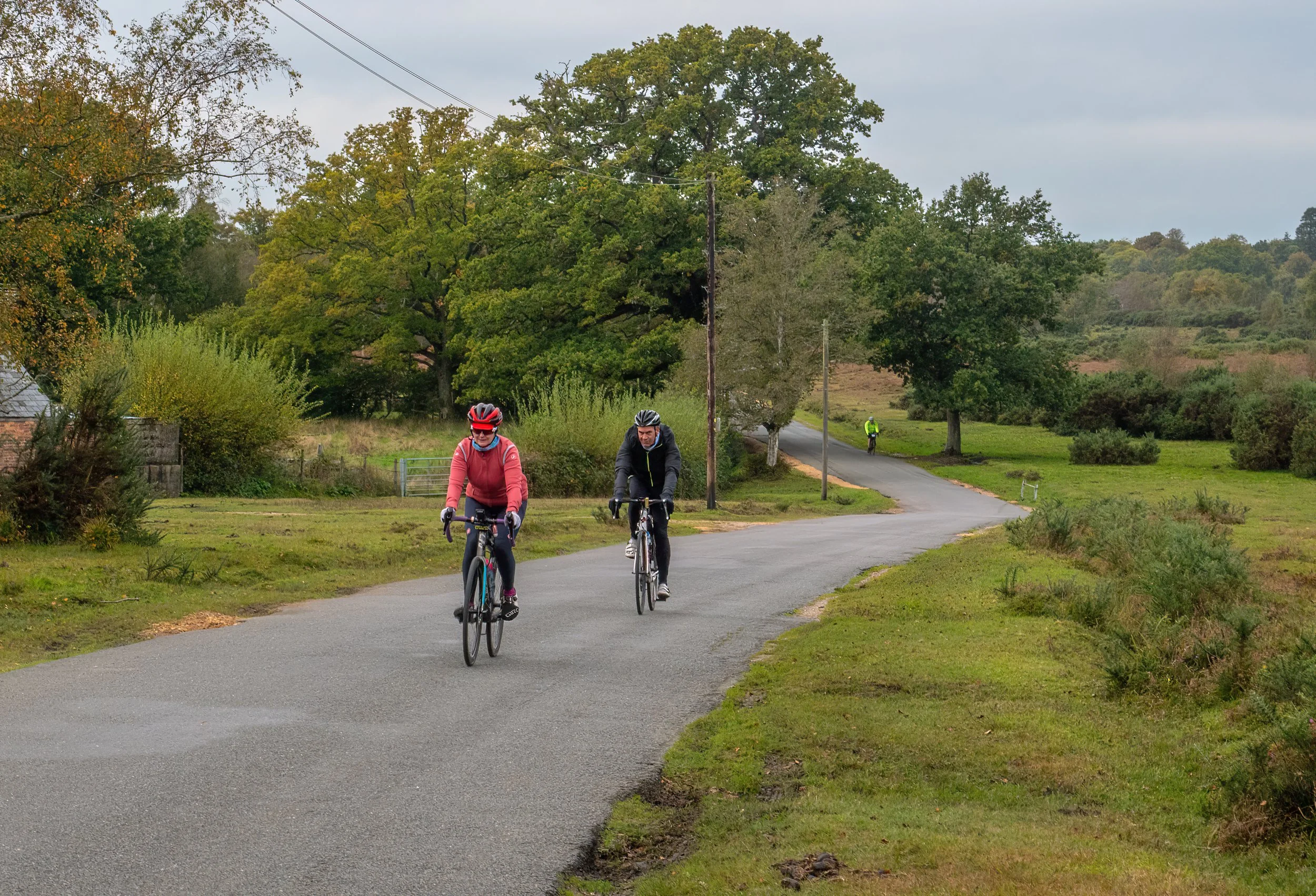 Two people riding bicycles on a winding country road surrounded by green trees and grass, with a utility pole and another person walking in the background on an overcast day.