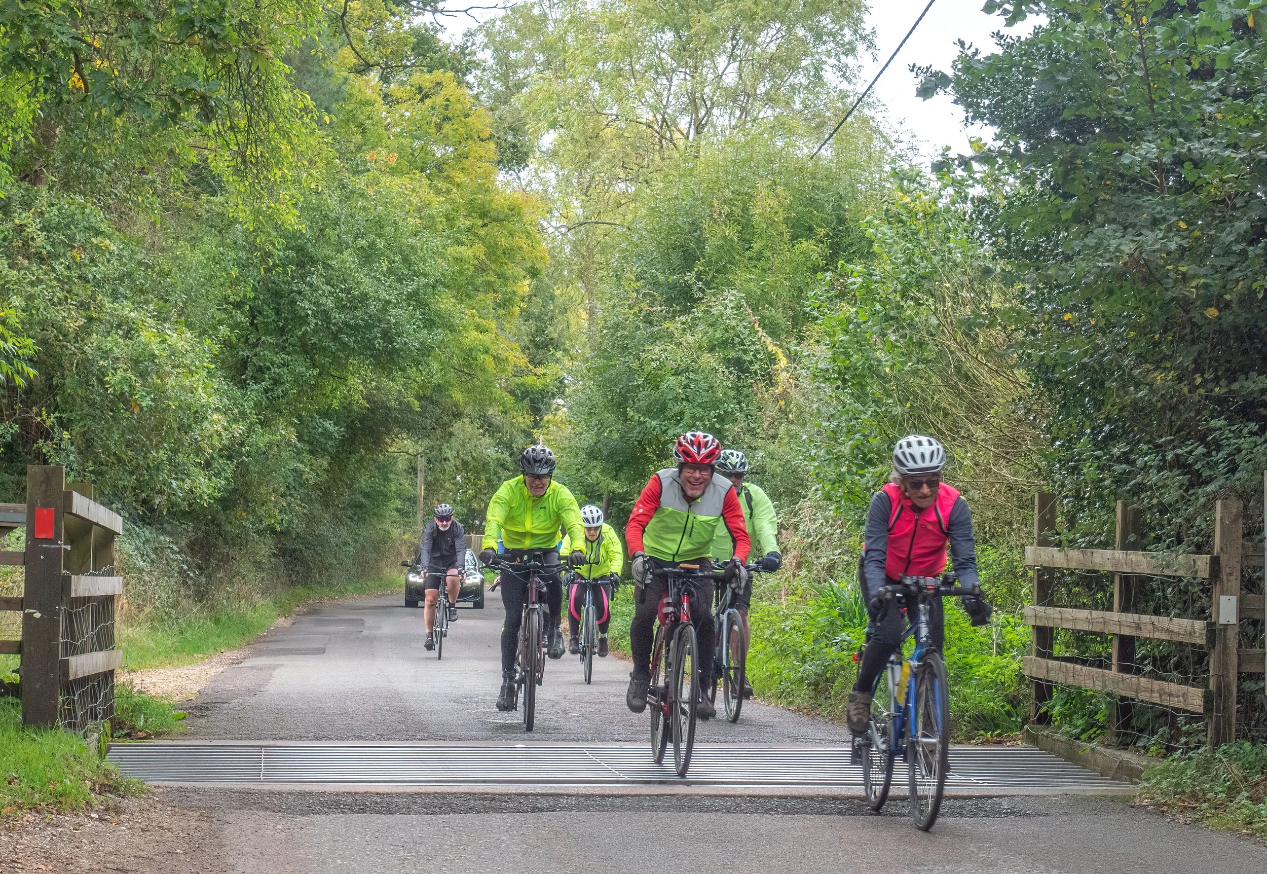 Group of cyclists wearing helmets and colorful jackets riding through a green, wooded area on a rural trail.