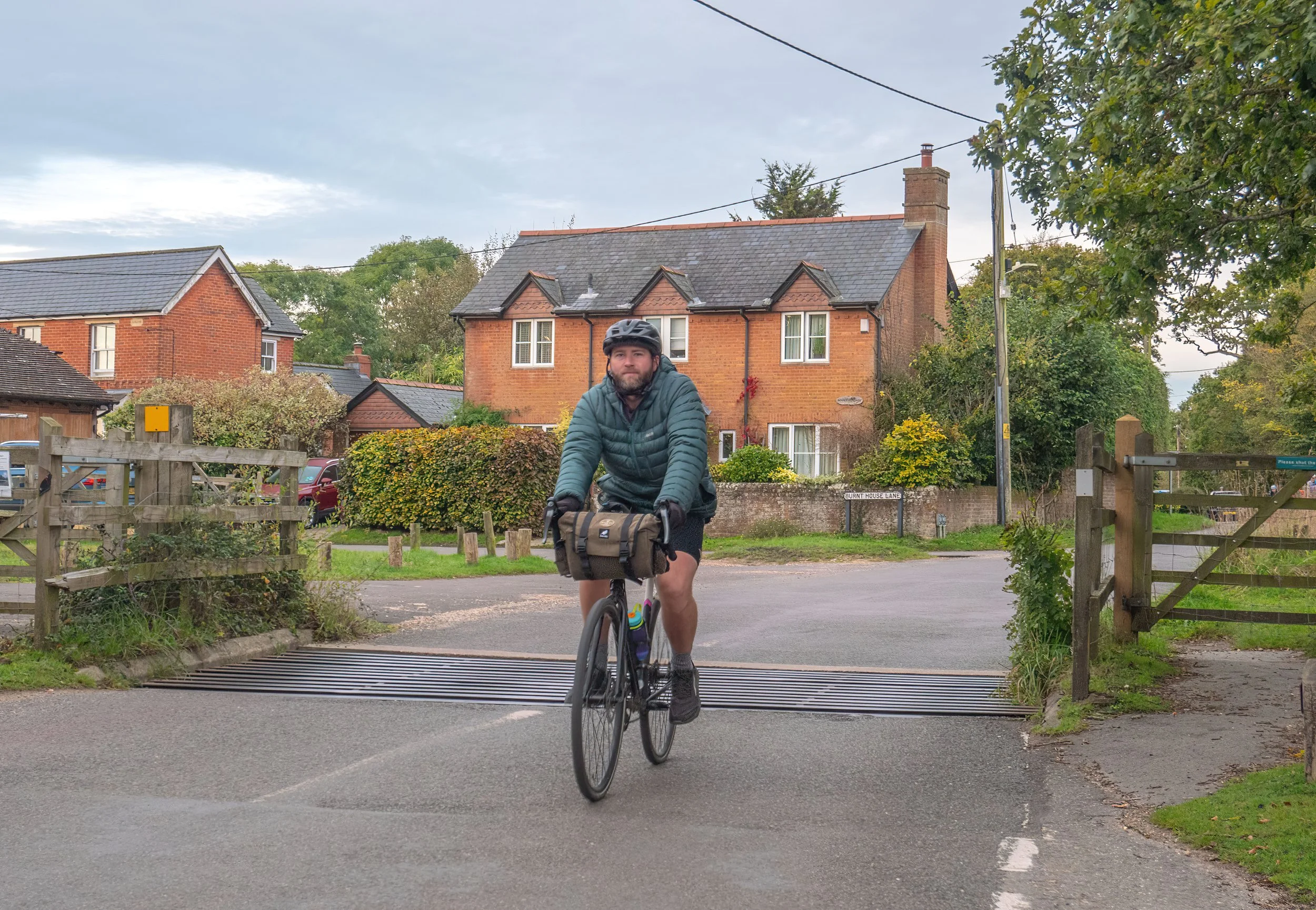 A man riding a bicycle crossing a speed bump in a residential neighborhood, holding the handlebars. He is wearing a helmet, a teal jacket, and sports a beard. The background shows brick houses, some with dark roofs, trees, and a cloudy sky.