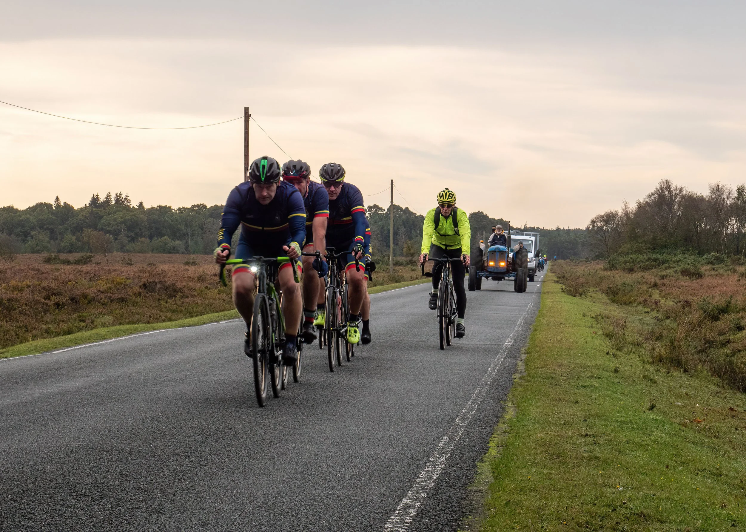 Group of cyclists riding on a rural two-lane road with a tractor following behind, surrounded by fields and trees under an overcast sky.